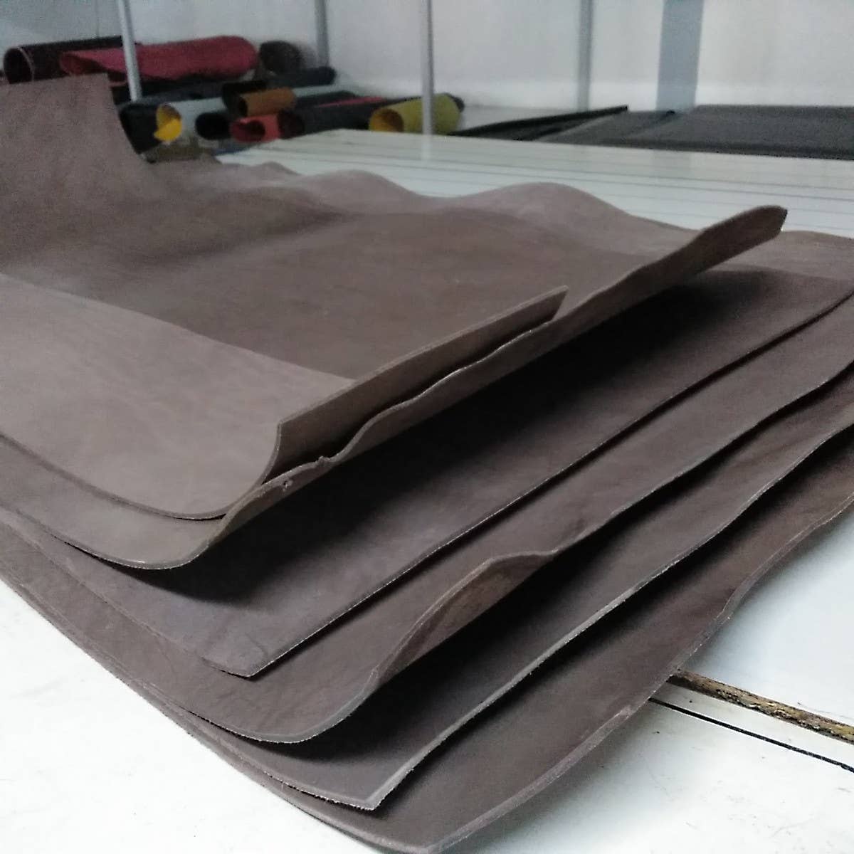 A Handcrafted Nubuck Leather Belt 3.5 cm by Creaciones Corpas is shown in a workshop, with flat sheets of brown Nubuck leather and rolled rubber sheets visible in the background.