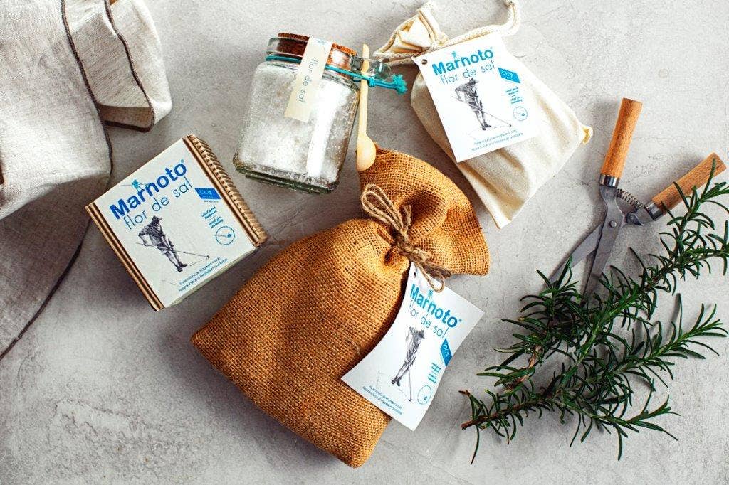 A Necton Salt Flakes in Cotton Bag 125g is displayed alongside scissors, a rosemary sprig, fabric bags, and Mar Marnoto Flor de Sal packets on a light stone surface with a cloth in the corner.