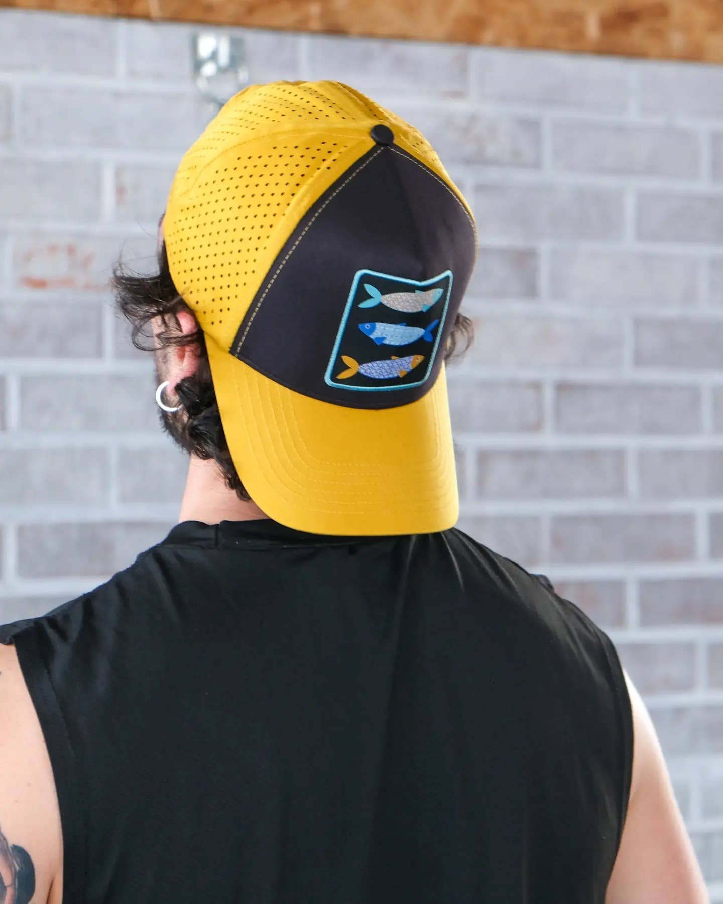 A person in a sleeveless black shirt and a Rodilha Cap-Sardines and Tiles (Yellow and Black) stands facing a light gray brick wall.