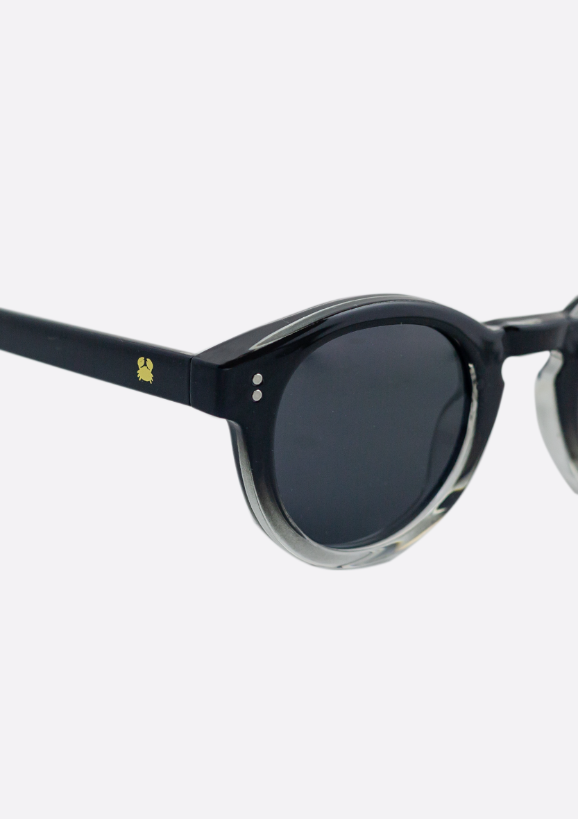 Close-up of Shiny Black Rodea Sunglasses by Oceanides, featuring dark polarized lenses and a small yellow turtle logo on the temple, crafted from recycled plastic, set against a white background.