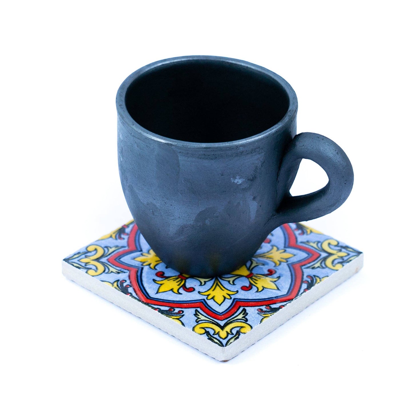 A black ceramic mug rests on a Tile-Inspired Ceramic & Cork Coaster from Meninas Bonitas Cork, featuring a decorative red, yellow, and blue pattern on a square base, set against a white background.