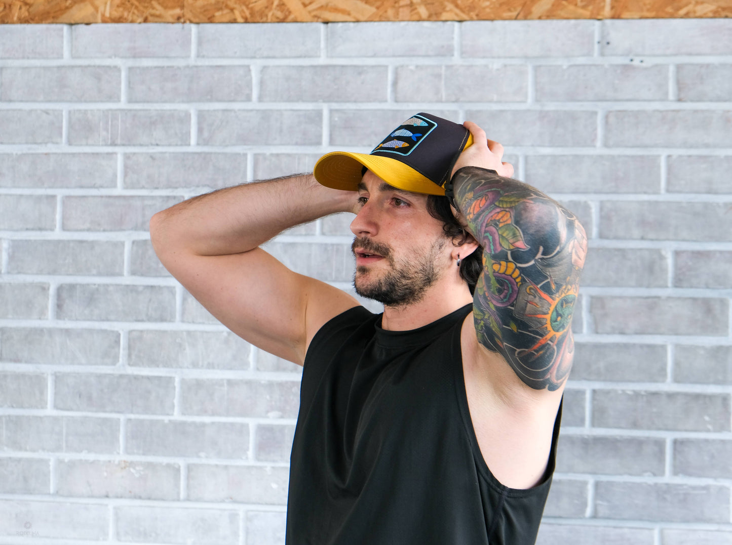 A man with a colorful arm tattoo and black sleeveless shirt stands in front of a light gray brick wall, adjusting his Rodilha Cap-Sardines and Tiles (Yellow and Black) with both hands, looking ahead.