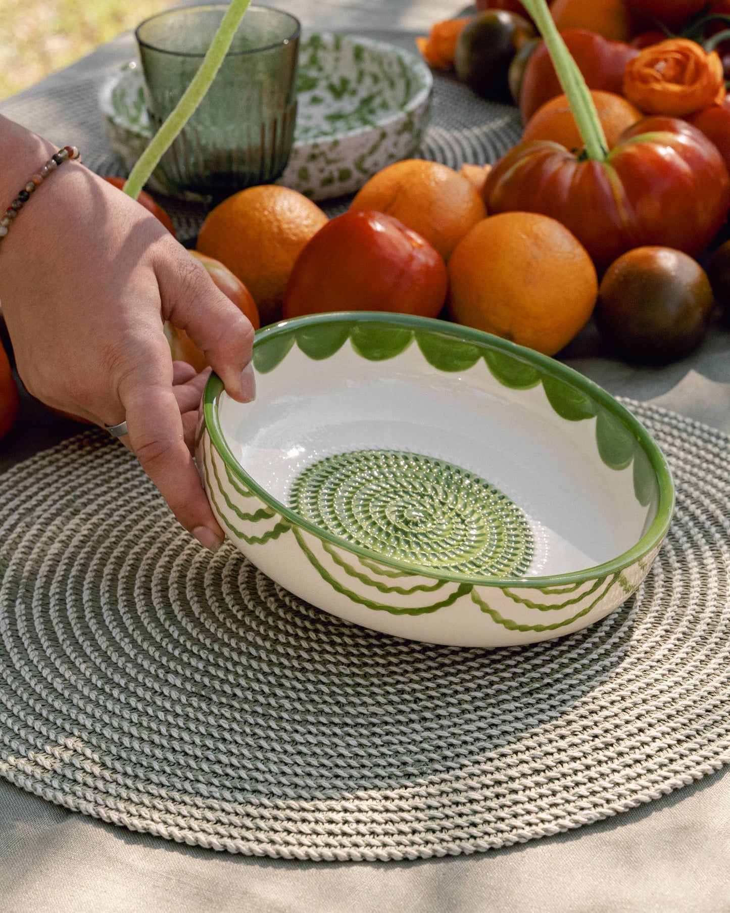A hand places the Iberica Large Pottery Grater Plate - Roots, a decorative green-and-white handcrafted bowl, on a woven placemat amid oranges, tomatoes, a glass, and another patterned bowl on the table.