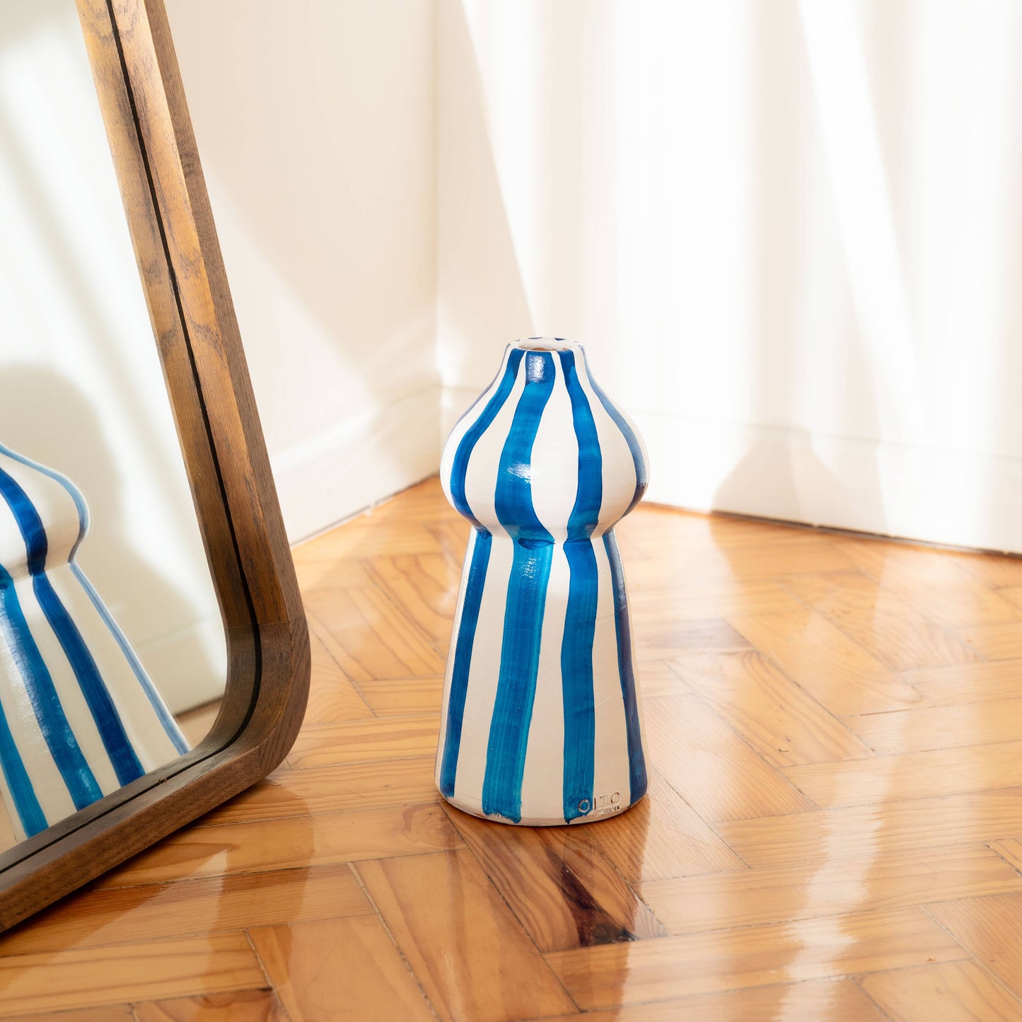 The Lucca vase by OITO Atelier, featuring blue and white stripes, is handcrafted and placed on a wooden floor beside a large mirror that reflects its design and the room’s bright natural light.