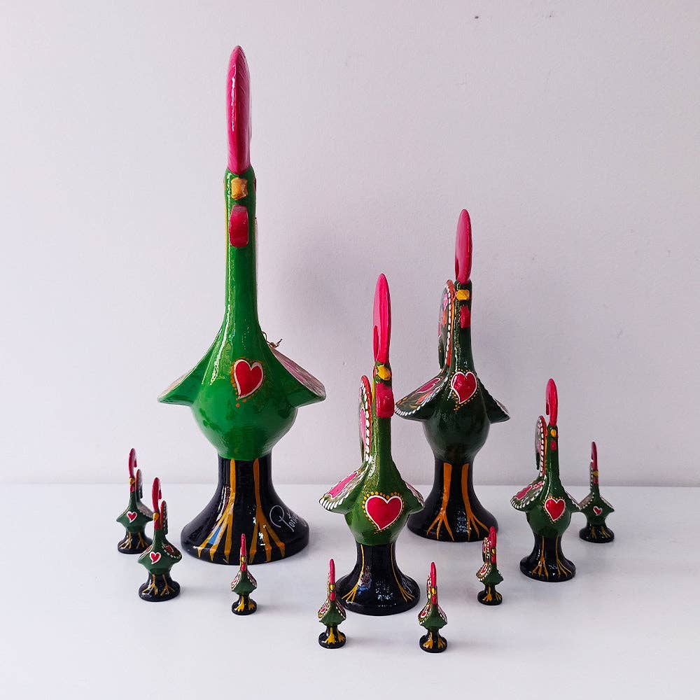 A group of Ibericas Barcelos Rooster figurines—mainly green with red crests and hearts—are arranged on a white surface, reflecting Portuguese art and the iconic design by Pretty things from Portugal.