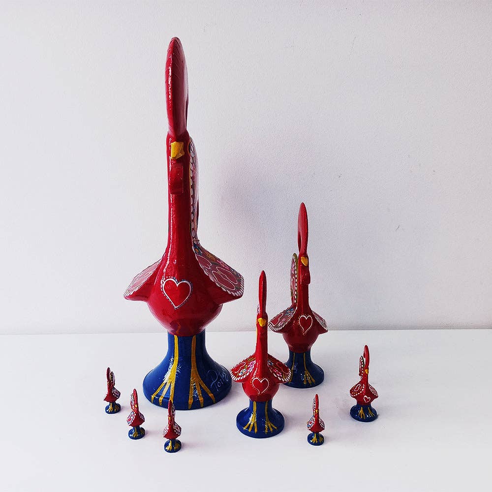 Nine Barcelos Rooster figurines by Iberica - Pretty things from Portugal, in red and blue and various sizes, are arranged on a white surface with the largest in the center and smaller ones surrounding it, set against a plain white background.