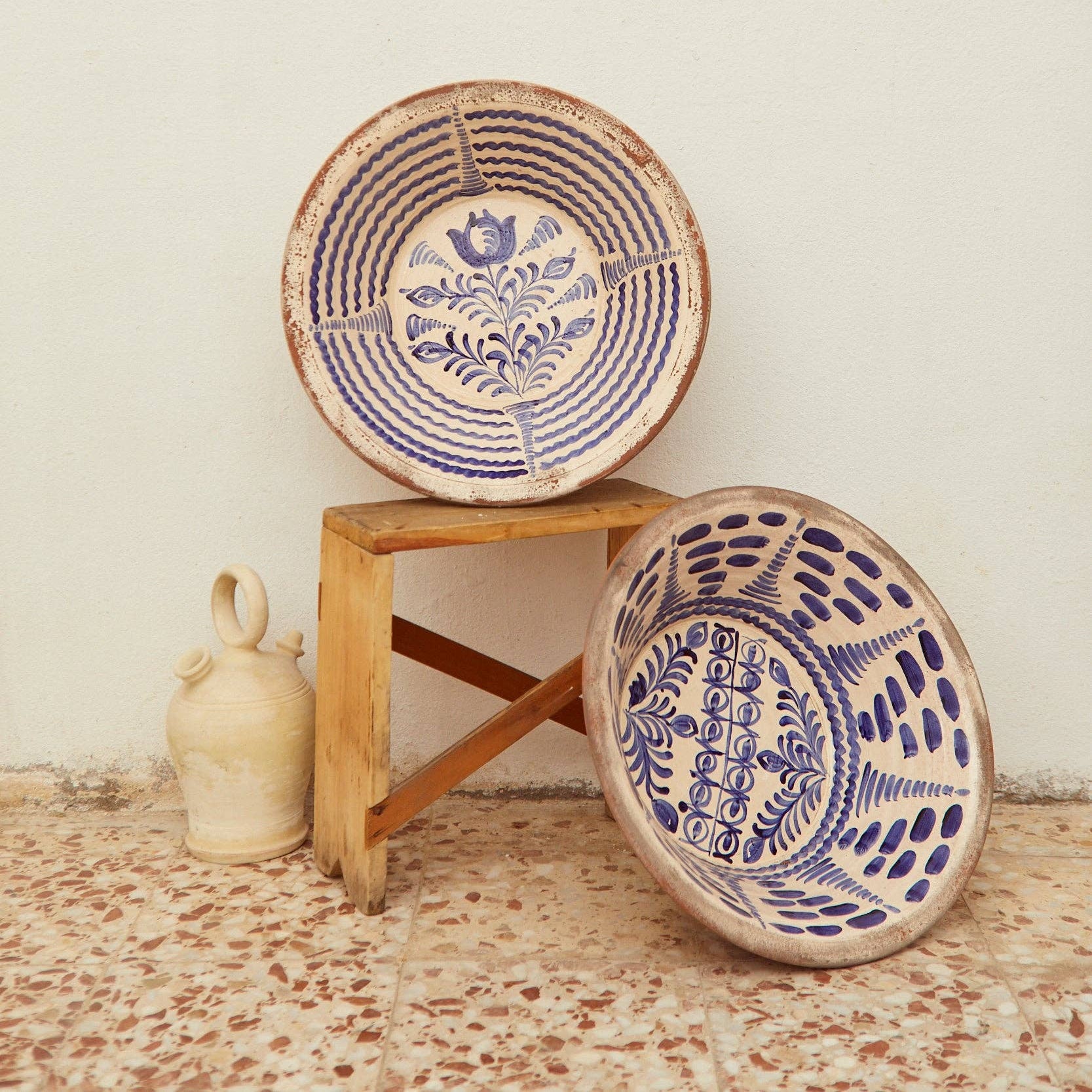Two large Blue Lebrillos Granaínos Bowls by Eturel Madrid, featuring blue patterns, sit on and beside a small wooden stool. A pale jug stands in the background, evoking Andalusian pottery against a plain wall and tiled floor.
