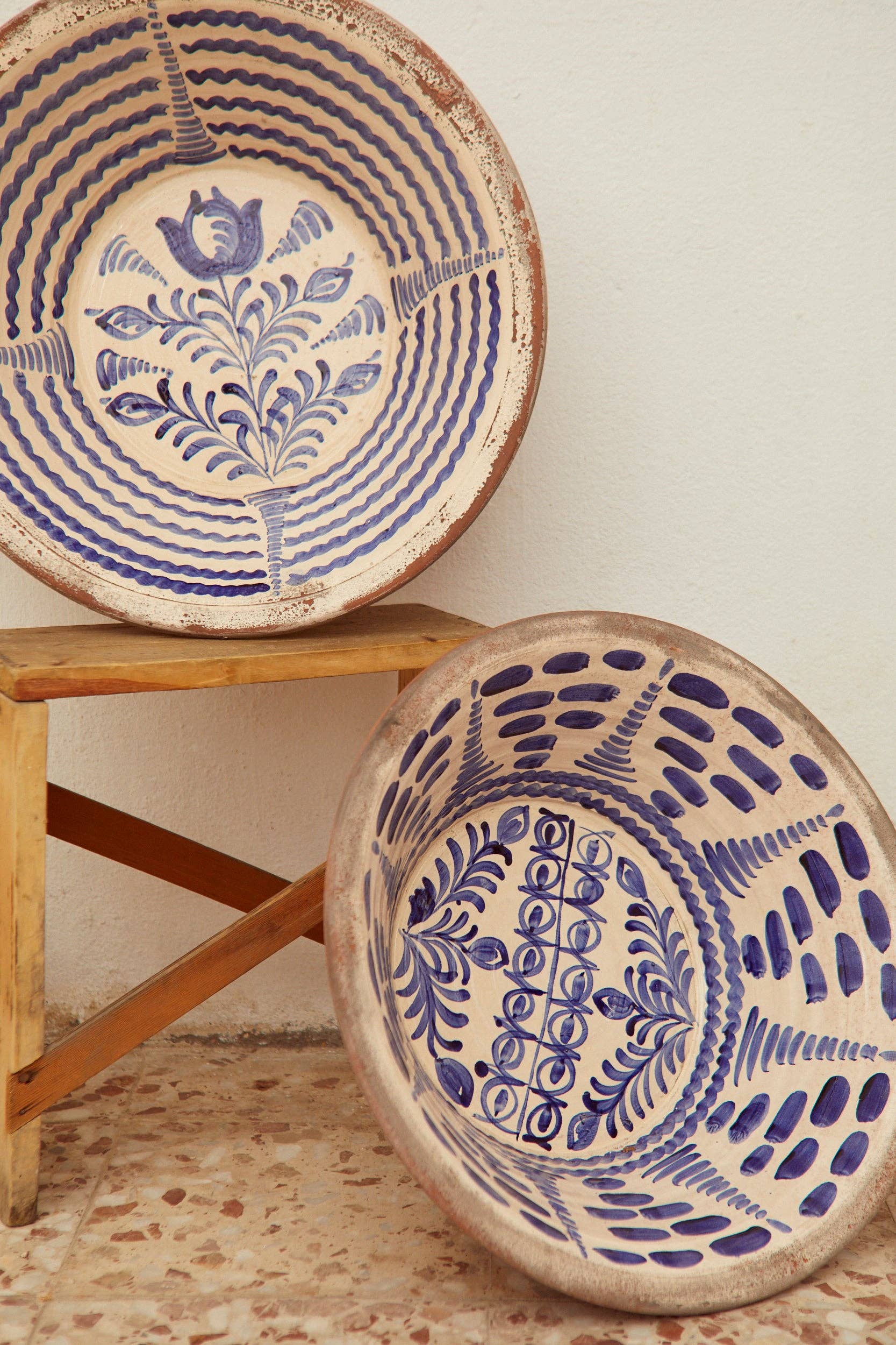 Two large Blue Lebrillos Granaínos Bowls by Eturel Madrid, featuring intricate blue floral and geometric designs, are displayed—one on a wooden stool and the other resting against it on a tiled floor against a white wall.