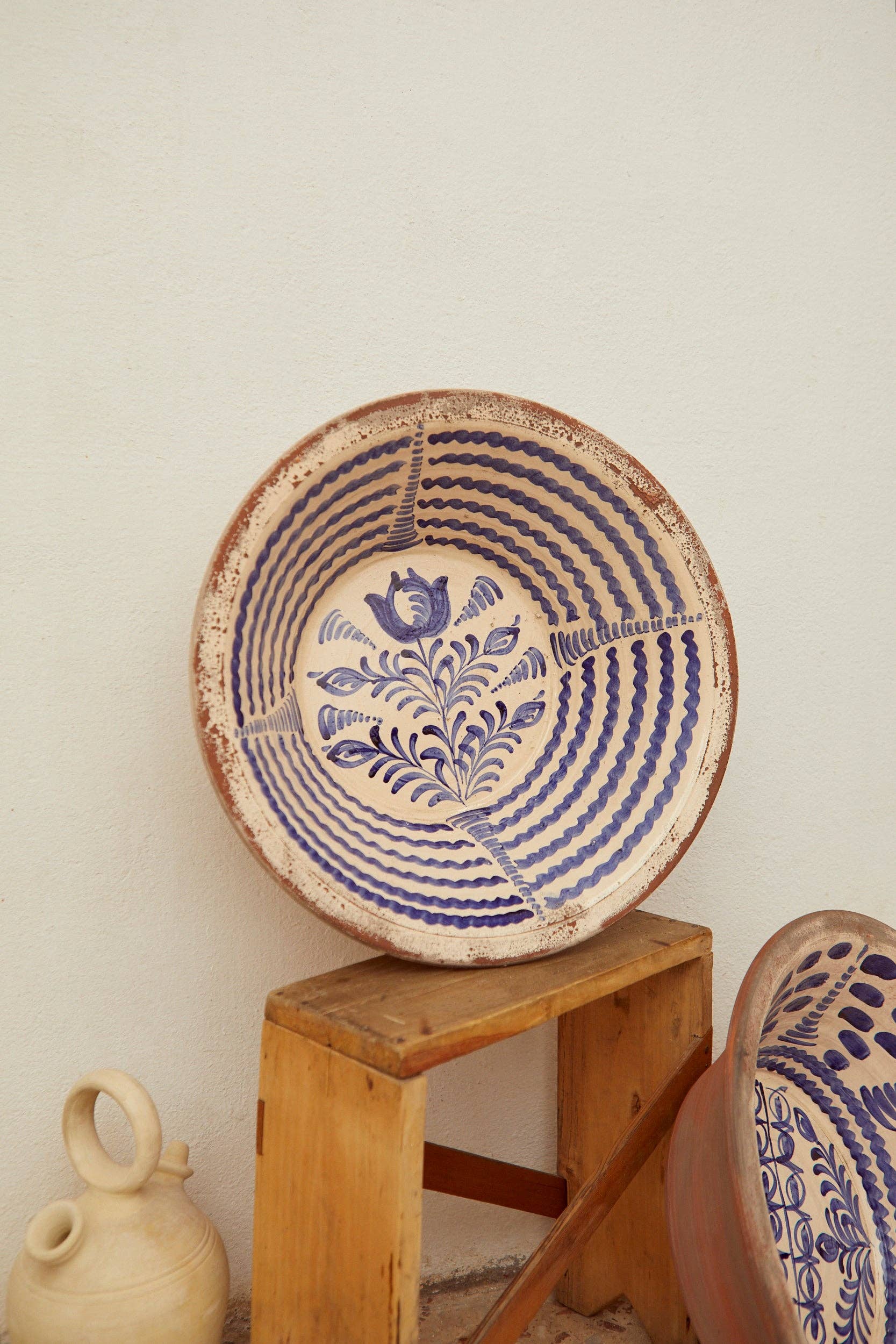 The Blue Lebrillos Granaínos Bowl by Eturel Madrid, featuring blue floral and geometric designs, rests on a wooden stool near a white wall; below, a smaller jug and an Andalusian-style bowl are partly visible.