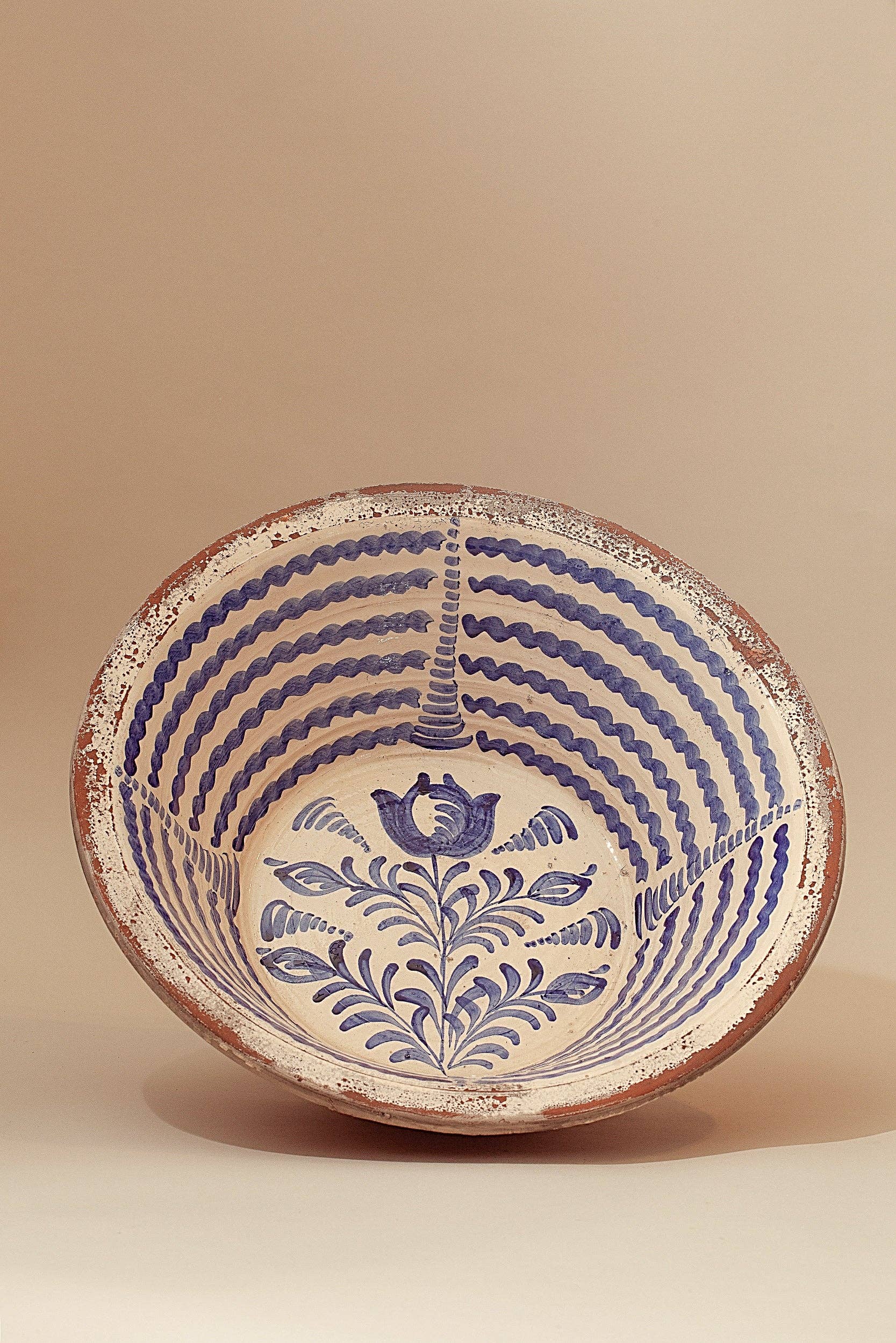 The Blue Lebrillos Granaínos Bowl by Eturel Madrid is a handcrafted Andalusian ceramic piece with a vintage finish, featuring a white interior with blue wavy lines, a central floral motif, and a rustic, worn rim for added character.