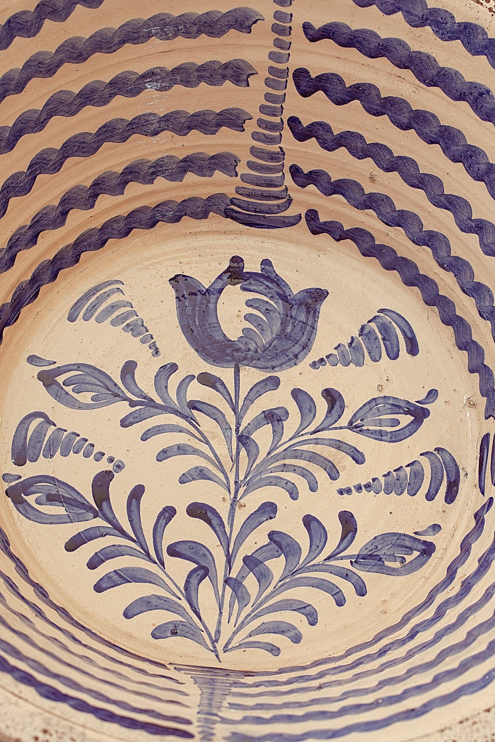 A close-up of the Blue Lebrillos Granaínos Bowl by Eturel Madrid, showcasing blue hand-painted wavy lines on the rim and a stylized blue floral center, inspired by Andalusian pottery, set against a light beige background.