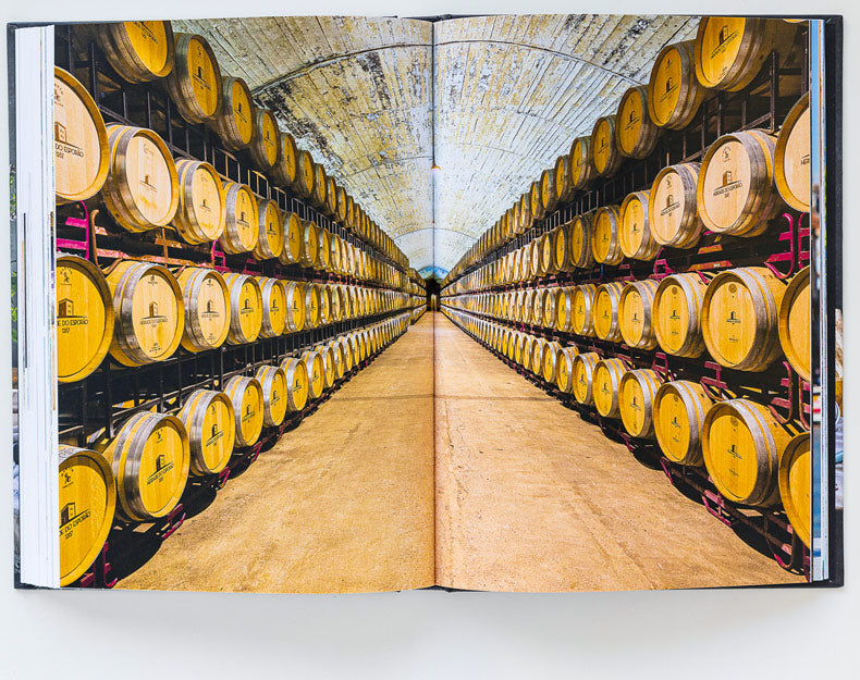 Photo of an open book reveals a cellar lined with wooden wine barrels, capturing the spirit of Bread, Olive Oil & Wine by ObjectoAnonimo and evoking Mediterranean flavors beneath an arched ceiling.