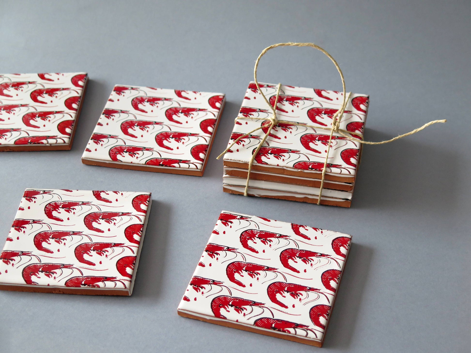 Gamba Design Ceramic Coasters by Bussoga, featuring a red and white abstract wave pattern, are displayed on a gray background—four stacked and tied with twine, three laid flat. Handmade in Spain.