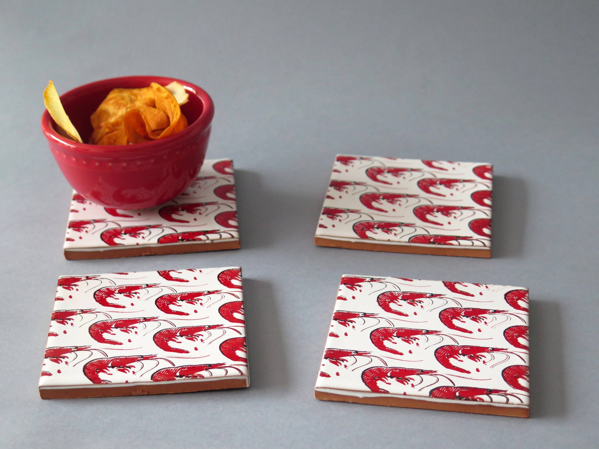 A small red bowl with chips sits on a Bussoga Gamba Design Ceramic Coaster featuring a red shrimp pattern. Three matching coasters are nearby on a gray surface. All coasters are handmade in Spain.