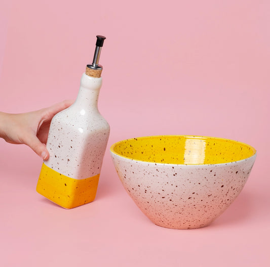 A hand holds the CANELA Oil and Salad Bowl from Alfar Tierra Cocida, a speckled white and yellow handmade salad kit, set against a pink background.