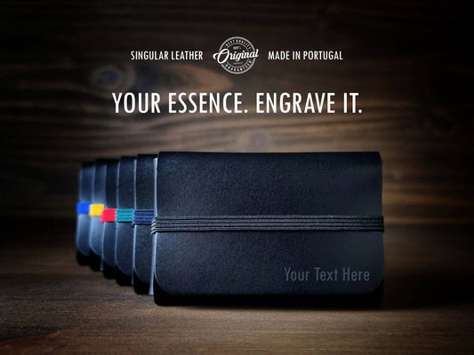 A row of Singular Leather Carbon Black Leather Wallets with colored bands is displayed on wood. The text above reads: “Your Essence. Engrave It. Singular Leather. Made in Portugal.” Now includes RFID protection for your security.