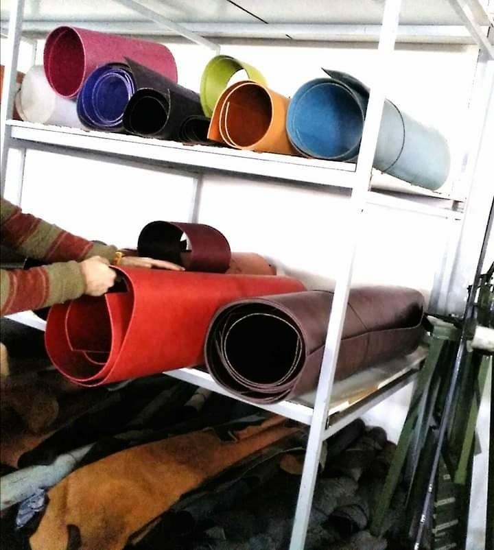 Several rolls of colorful leather, ideal for handmade items like the Castilian Handmade Leather Belt 3.5 cm by Creaciones Corpas, are stored on metal shelves as someone arranges a large red-brown roll; darker hides are stacked below.