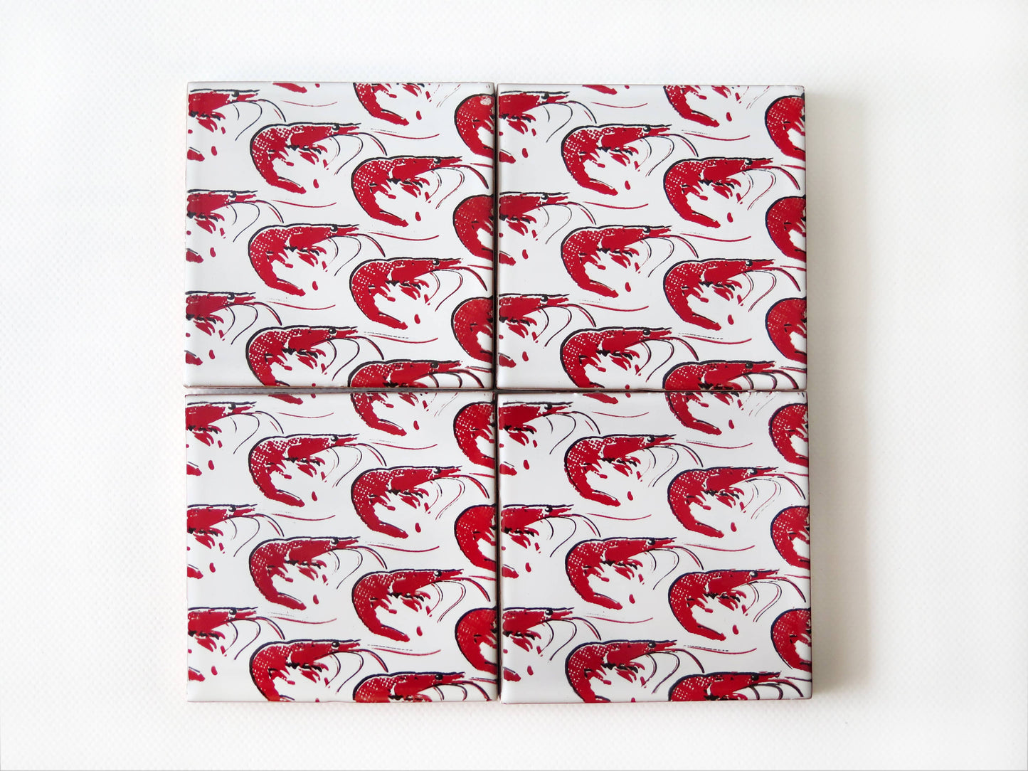 Four Gamba Design Ceramic Coasters by Bussoga, handmade in Spain, are wrapped in white paper featuring a red shrimp pattern and arranged in a tidy 2x2 grid on a white background.