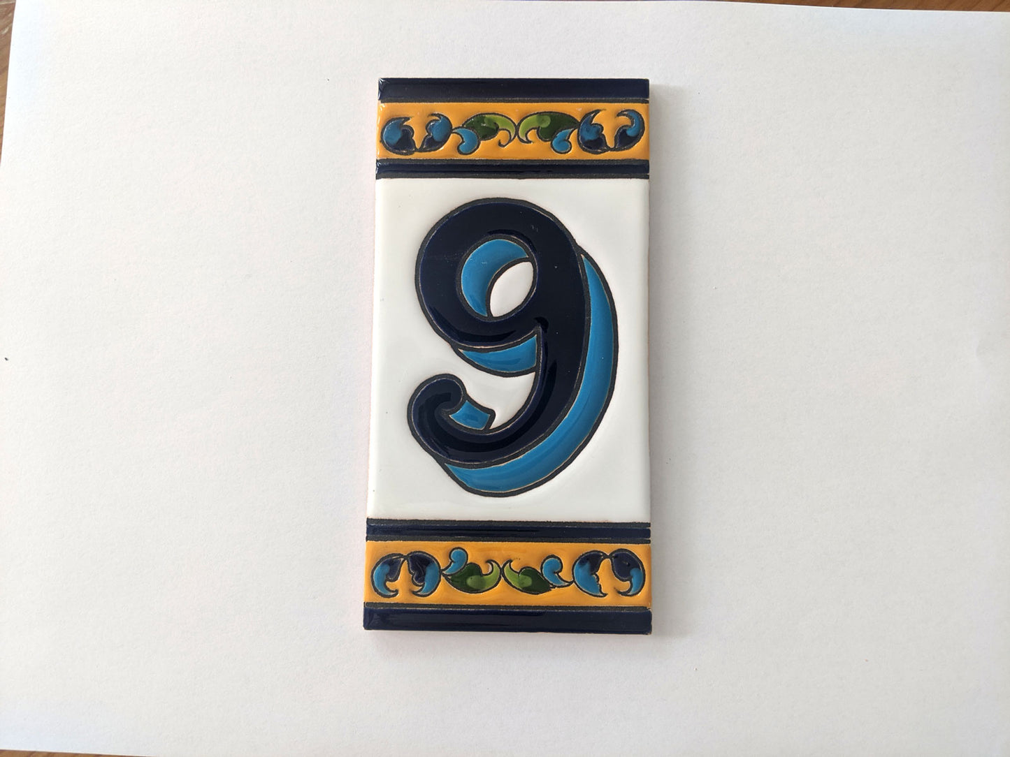 The Iberica Ceramic Number Tile 15cm features a bold blue number 9 on a white background, accented by handpainted yellow, blue, and green floral designs on the borders—perfect for adding charm to your house numbers.