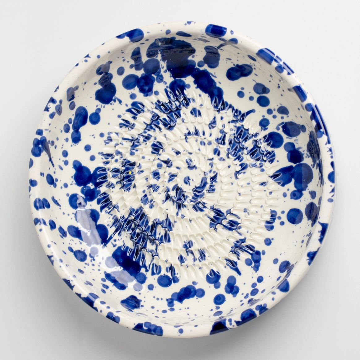 The Handmade cheese and vegetable grater plate - PERSIA by Tierra Cocida is a round ceramic plate with blue splatter on white, handmade in Spain, featuring a ridged center—ideal for Mediterranean kitchens.