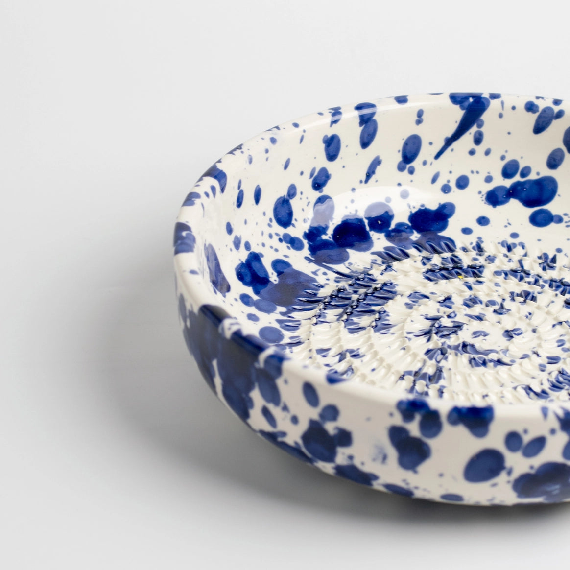 The Handmade cheese and vegetable grater plate - PERSIA by Tierra Cocida is a round, shallow white ceramic plate with blue splatter patterns and a ridged center, handmade in Spain—ideal for Mediterranean kitchen decor. Photographed on a plain light background.