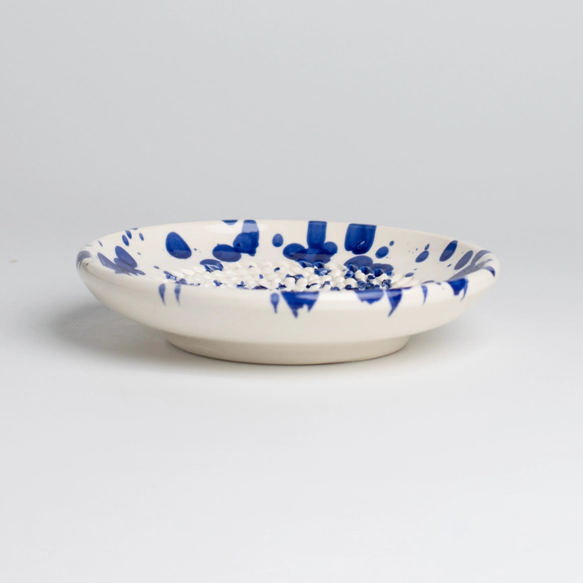 The Handmade cheese and vegetable grater plate - PERSIA by Tierra Cocida features a shallow, round ceramic design with a white base and blue splatter pattern—handmade in Spain, ideal for Mediterranean kitchen decor. Shown on a light gray background.