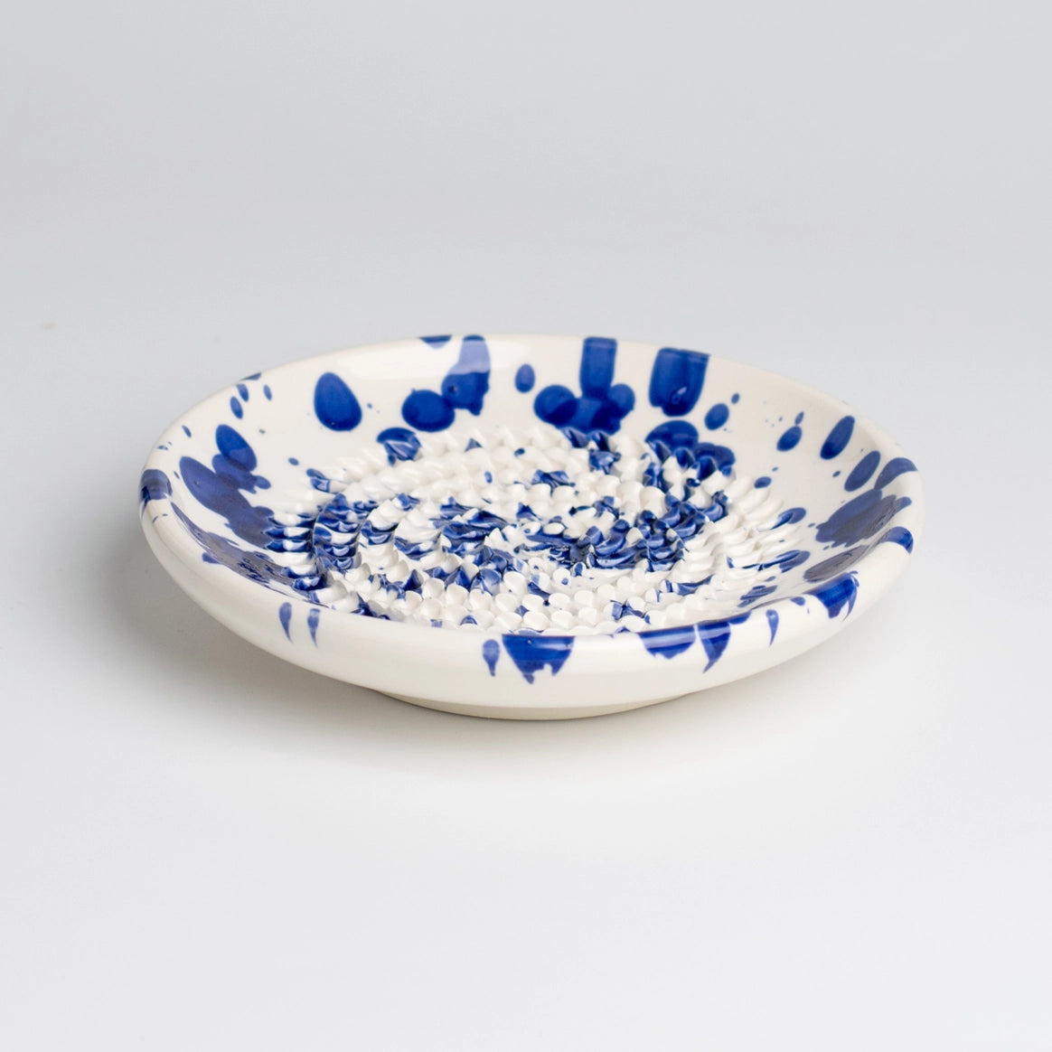 The Tierra Cocida Handmade cheese and vegetable grater plate - PERSIA features a white ceramic base with blue splatter, raised center spikes for grating, and Mediterranean style. Handmade in Spain—ideal as functional kitchen decor.