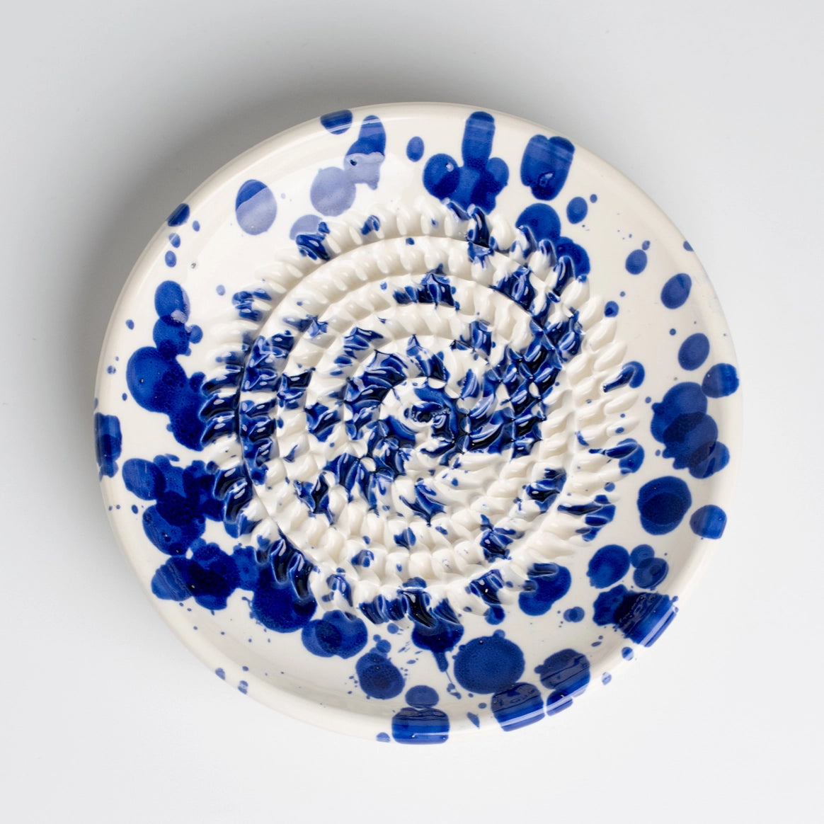 The Tierra Cocida Handmade cheese and vegetable grater plate - PERSIA is a round white ceramic plate, handmade in Spain with a textured spiral center and bold blue splatters. Perfect for Mediterranean kitchens. Background is plain light gray.