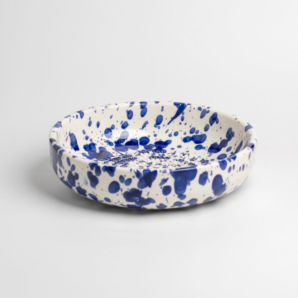The Handmade cheese and vegetable grater plate - PERSIA by Tierra Cocida boasts a white base with bold blue splatter, ideal for Mediterranean kitchen decor. Expertly handmade in Spain, it’s shown against a plain white background.