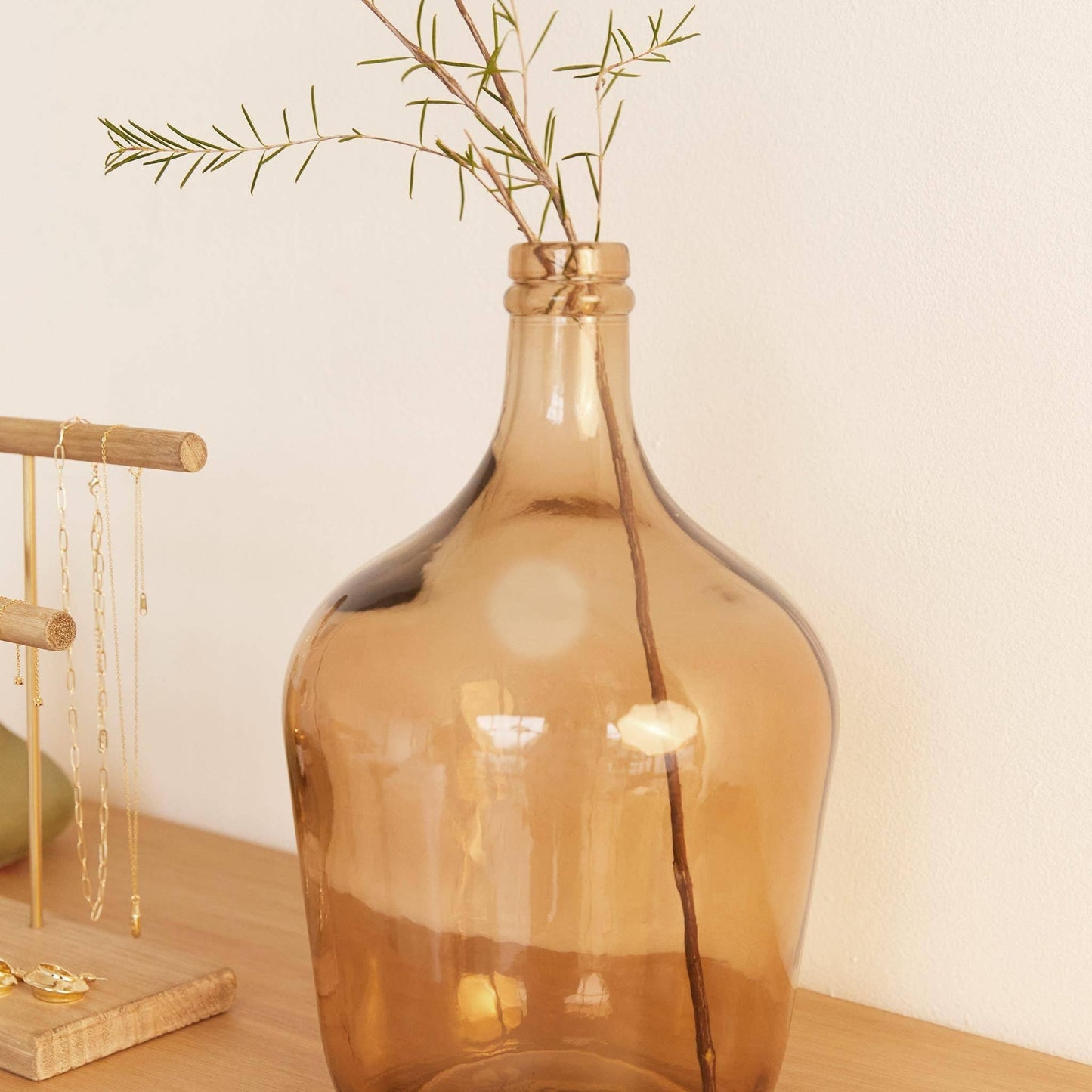 The Hannun Luiza Demijohn Vase, large and amber-colored with a narrow neck, sits on wood holding a thin branch with sparse leaves. Nearby, a wooden jewelry stand displays delicate necklaces.
