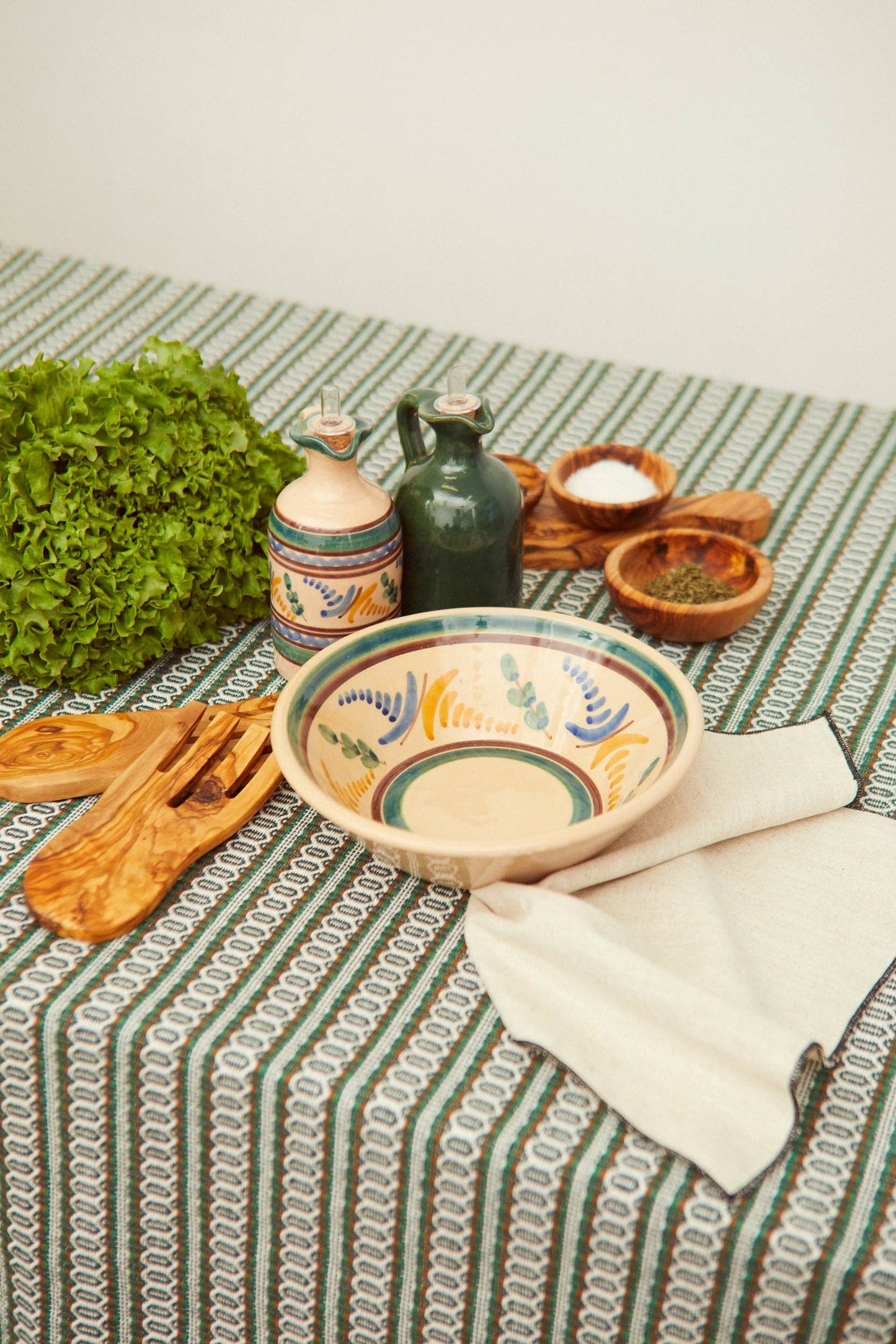 The Medina Oil Mill by Eturel Madrid, along with salt and spice bowls, wooden utensils, lettuce, and a folded napkin, rests on a green-and-white patterned tablecloth inspired by Andalusian ceramic tradition.
