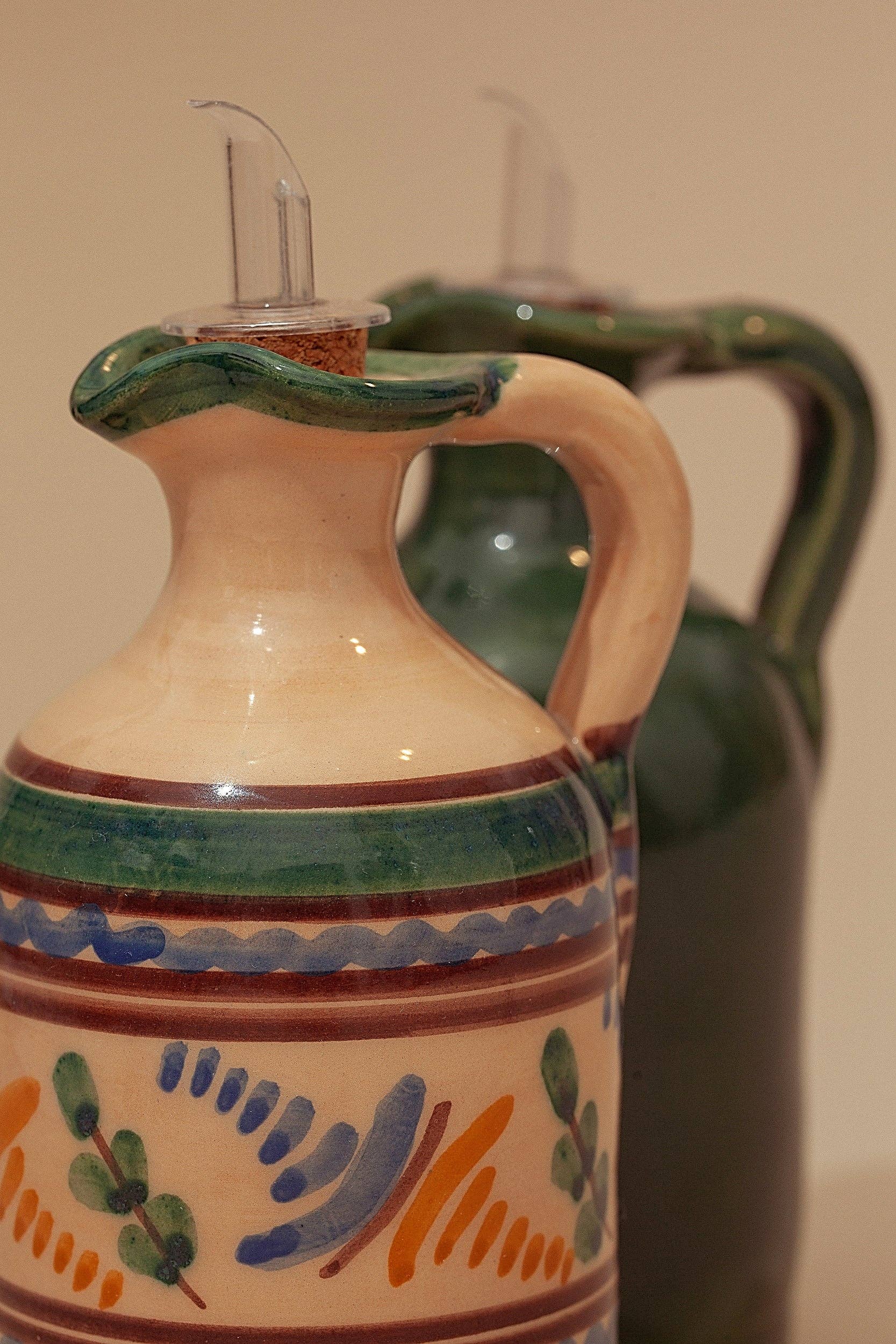 The Medina Oil Mill by Eturel Madrid features two dispensers: one in classic Andalusian ceramic with vibrant patterns and green details, the other in solid dark green. Both have handles and clear spouts for easy pouring.