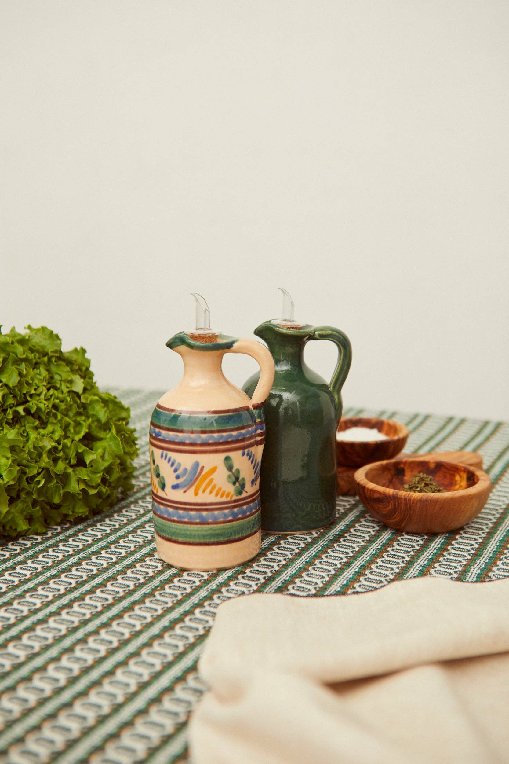Two Medina Oil Mill dispensers by Eturel Madrid—one with vibrant patterns, the other in solid green—are displayed on a patterned tablecloth next to fresh lettuce, small wooden bowls, and a beige napkin.