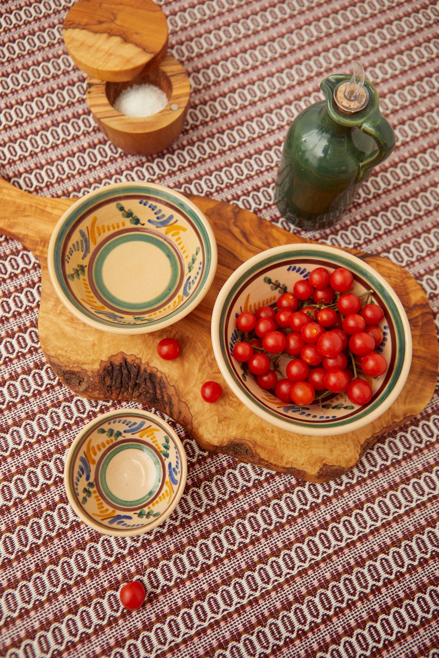 Three Eturel Madrid Medina Printed Ceramic Bowls—one with cherry tomatoes, two empty—sit on a wooden board atop a woven cloth. A small salt container and green glass oil bottle recall Córdoba heritage.