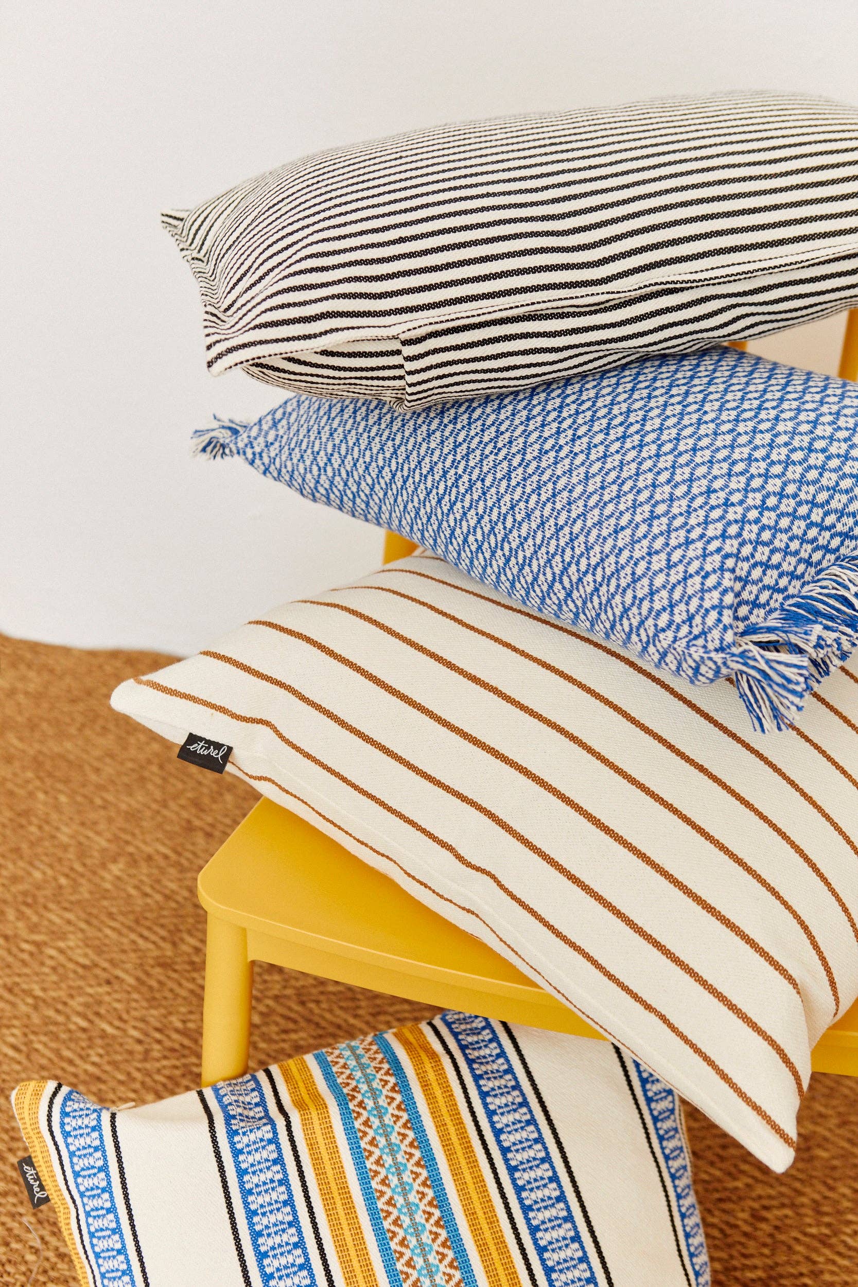 Four patterned pillows, including the Morellano Blue Woven Cushion Cover 50cmx50cm by Eturel Madrid, showcase striped and geometric designs in black, blue, and brown. They’re stacked on a yellow chair atop a textured brown woven rug.
