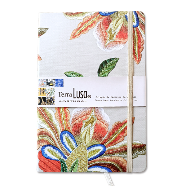 The Terra Lusa Castelo Branco Quilt Notebook features a white cover with colorful embroidered floral patterns inspired by Portuguese tradition, an elastic closure, and a paper band labeled Terra Lusa Portugal.