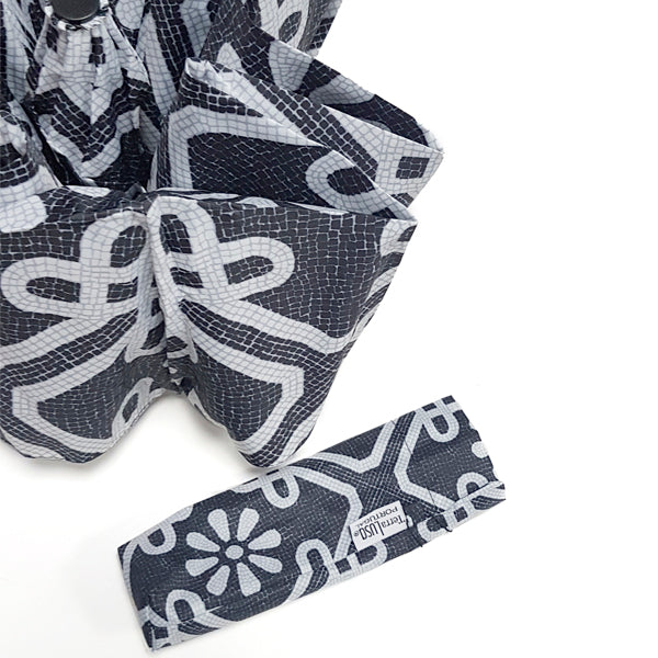A close-up of a black and white Sidewalk Umbrella by Terra Lusa, featuring geometric and floral patterns inspired by Portuguese sidewalks, shown with a matching “Urban Botanical” pouch.