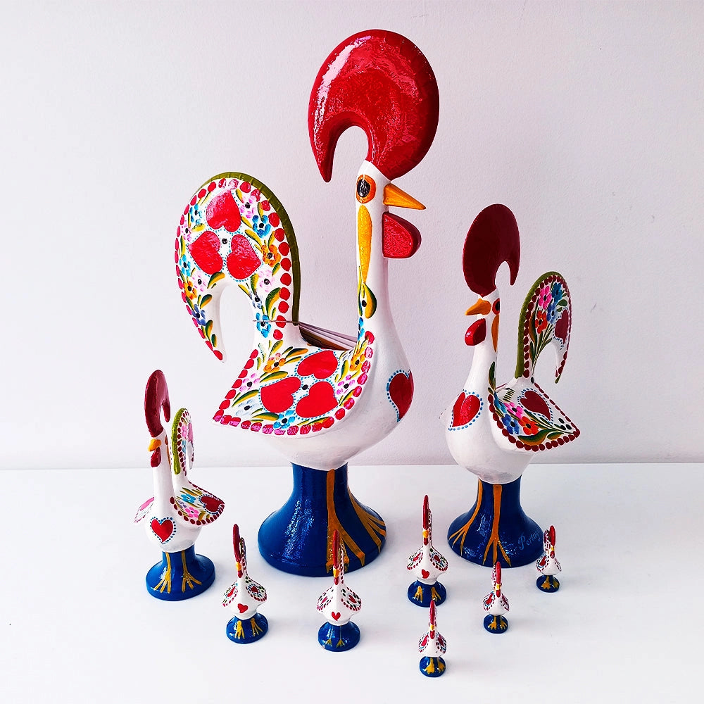 Nine colorful Barcelos Rooster ceramic figurines by Iberica - Pretty things from Portugal, in various sizes with bright red, yellow, green, and blue patterns, are displayed on a white surface against a plain background.