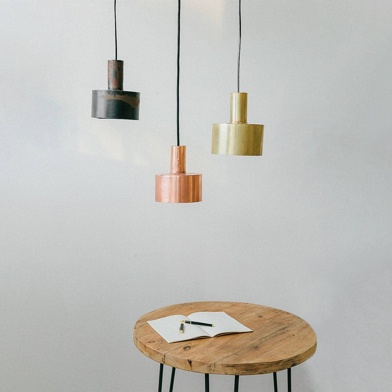 A round wooden table with an open notebook and pen sits beneath three Hannun Seber Lamps in black, gold, and copper, their hand-forged iron accents adding industrial style to a plain white wall.