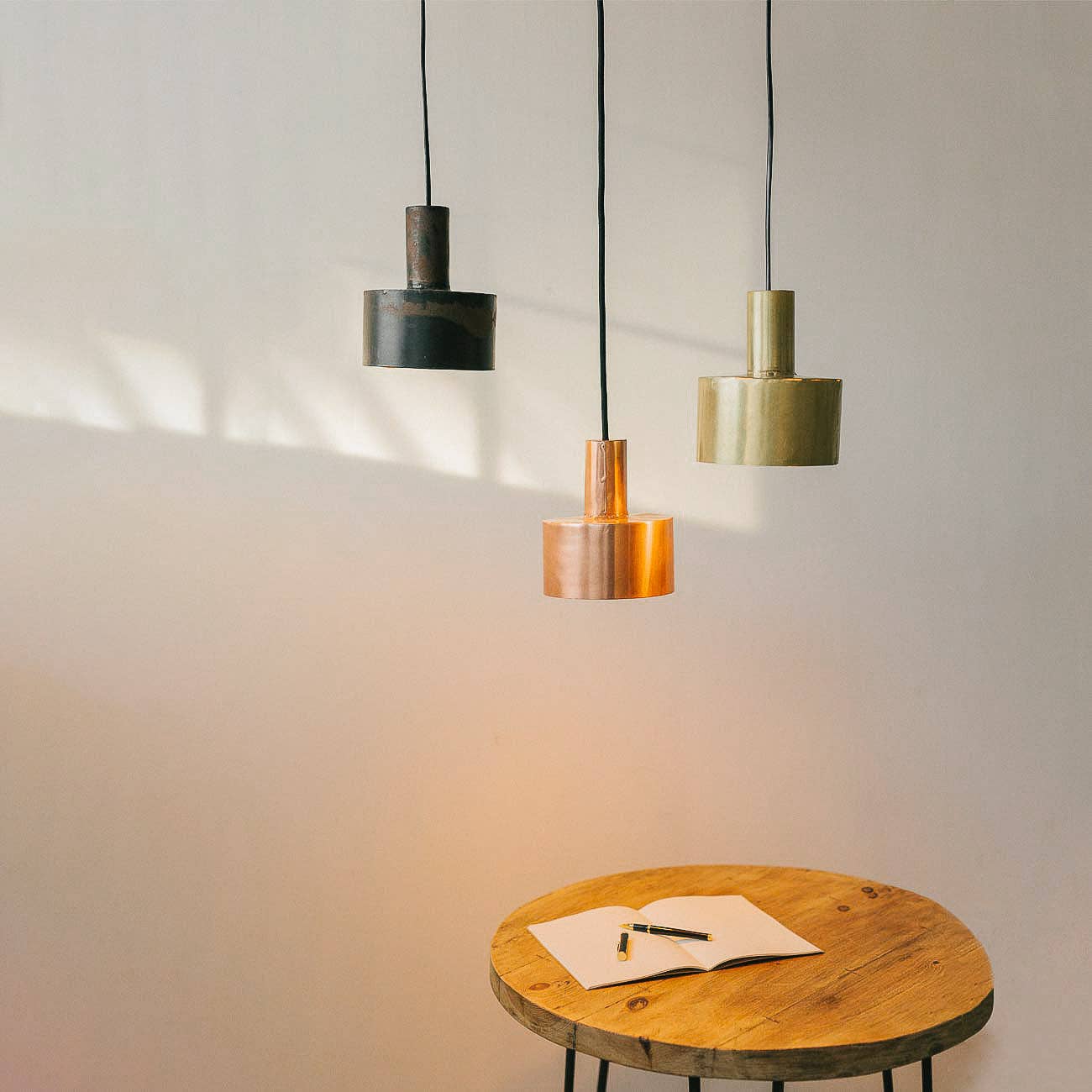 Three Seber Lamps by Hannun, available in 3 colours, hang above a round wooden table. An open notebook and pen rest below their glow, accentuating the softly lit, neutral wall.