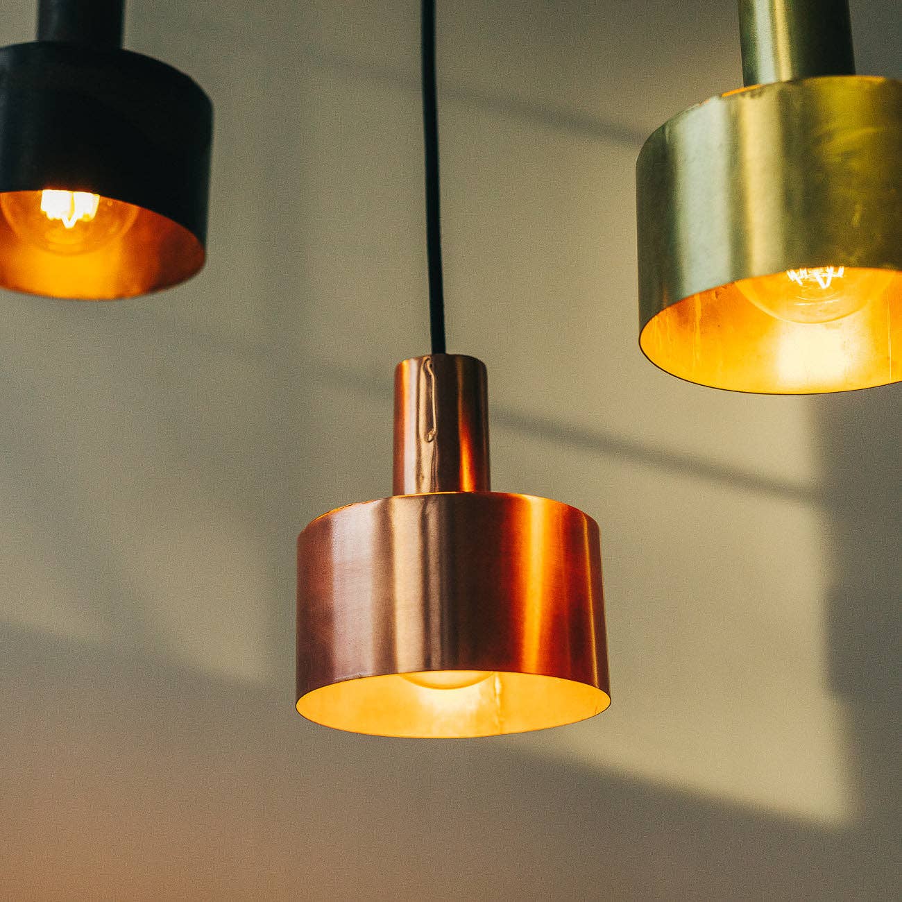 Three Seber Lamps by Hannun in black, bronze, and gold hang from the ceiling, their cylindrical shades casting a warm glow—perfect for industrial-style interiors.