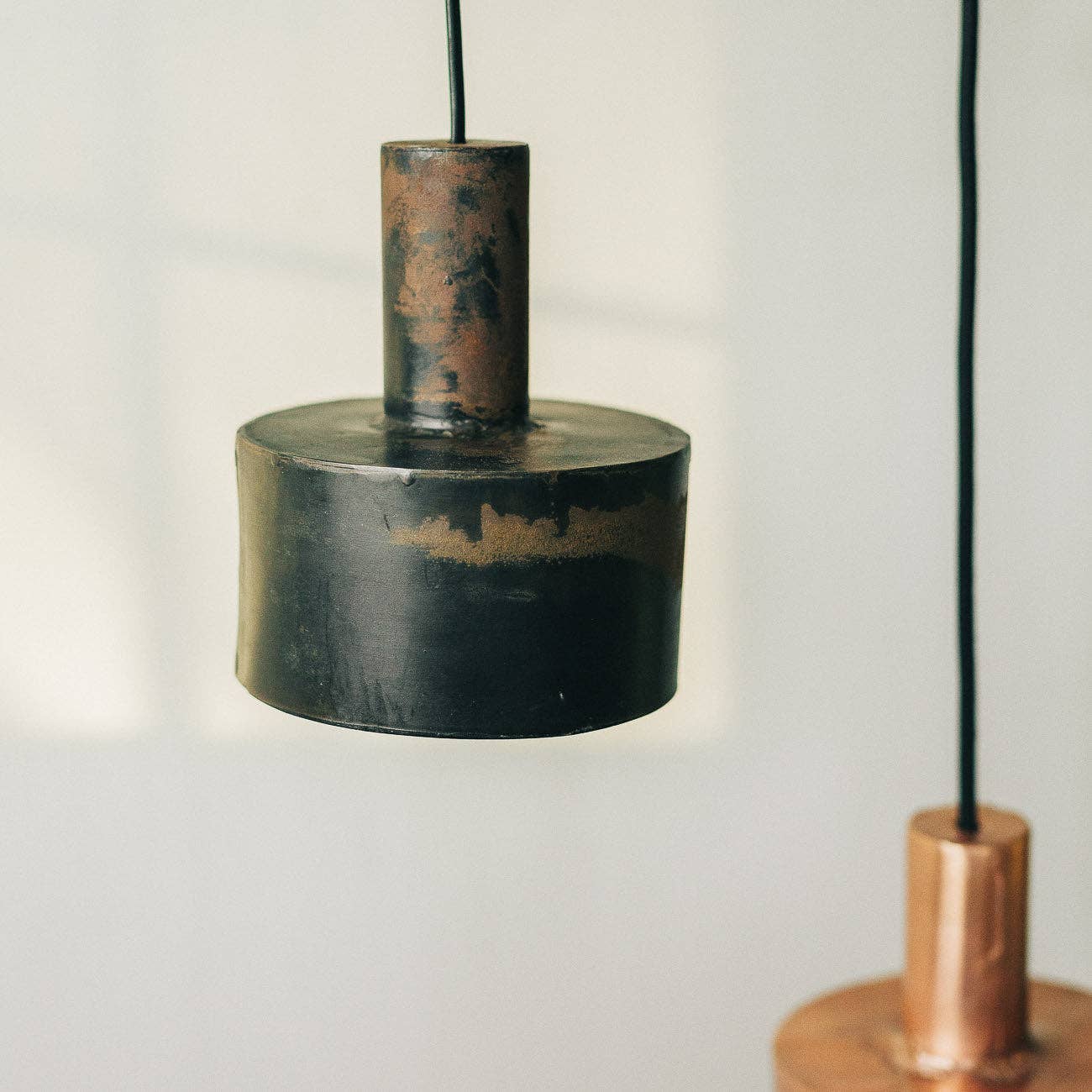 A close-up of two modern Seber Lamps by Hannun, featuring a rustic industrial design in hand-forged iron. Shown here in weathered black and copper tones, these pendant lights hang on black cords and are available in three colours.