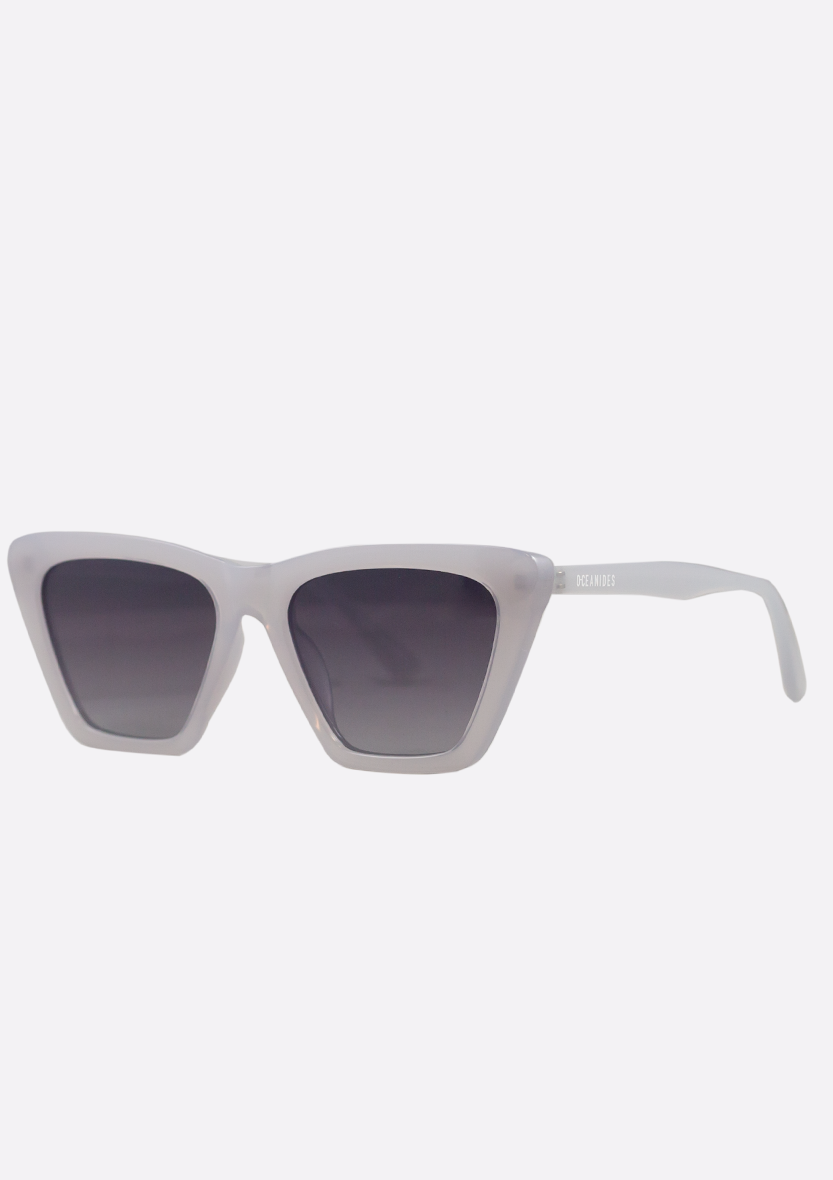 Oceanides Perse Crystal Baby Blue Polarized Sunglasses feature a translucent light gray cat-eye frame and dark tinted lenses, shown against a plain white background. Eco-friendly design.