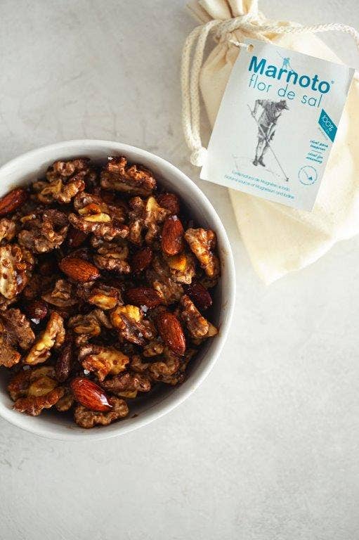 A white bowl with roasted walnuts and almonds sits on a light surface beside a cotton bag labeled Necton Salt Flakes 125g, perfect for adding a touch of delicate finishing salt.