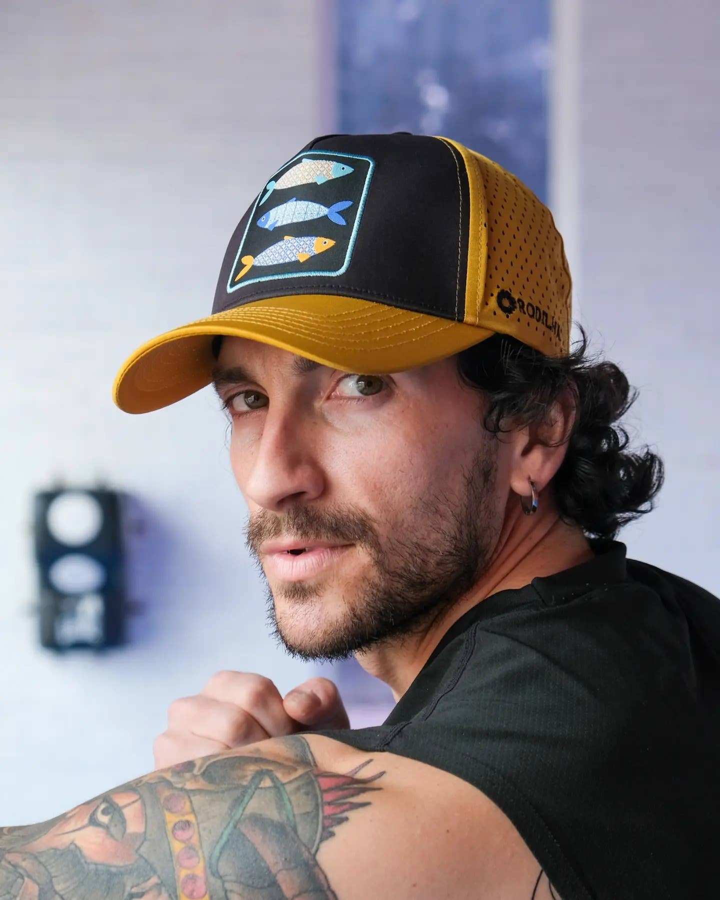 A man with curly hair and a beard wears the Rodilha Cap-Sardines and Tiles (Yellow and Black) featuring fish graphics. He has an earring, visible arm tattoos, and looks over his shoulder toward the camera indoors.