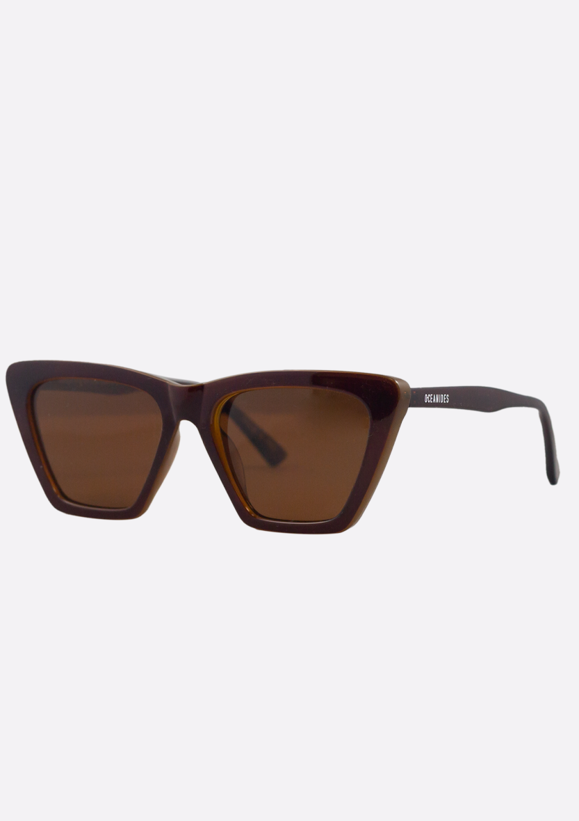 The Perse Double Brown Polarized cat-eye sunglasses by Oceanides feature thick dark brown frames and brown-tinted lenses, accented with a small white Oceanides logo on the left temple. Displayed on a light background, these are eco-friendly sunglasses.