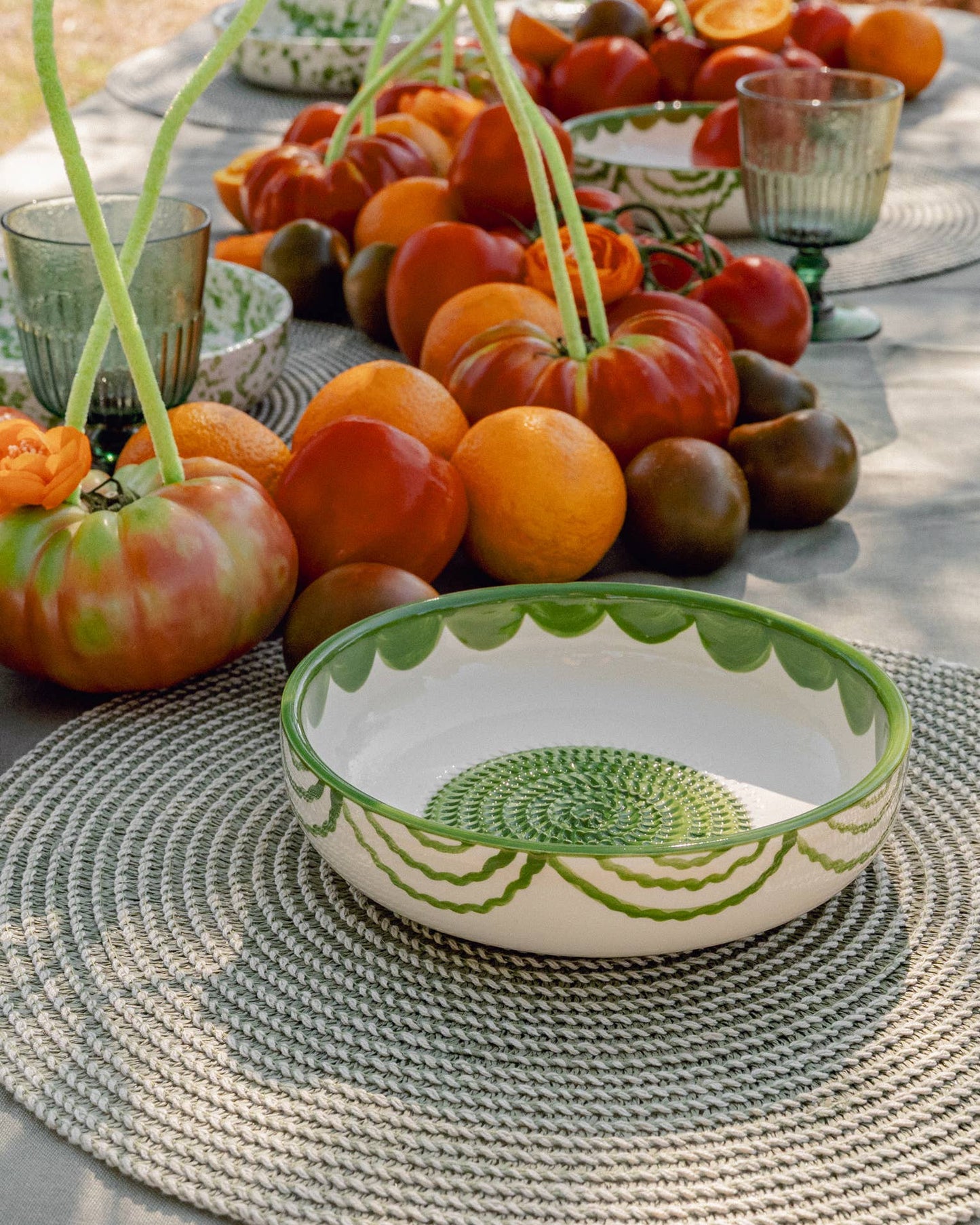A vibrant table setting features the Iberica Large Pottery Grater Plate - Roots, a handcrafted green and white bowl from Portugal, placed on a woven placemat with colorful tomatoes and glassware for a fresh, lively touch.