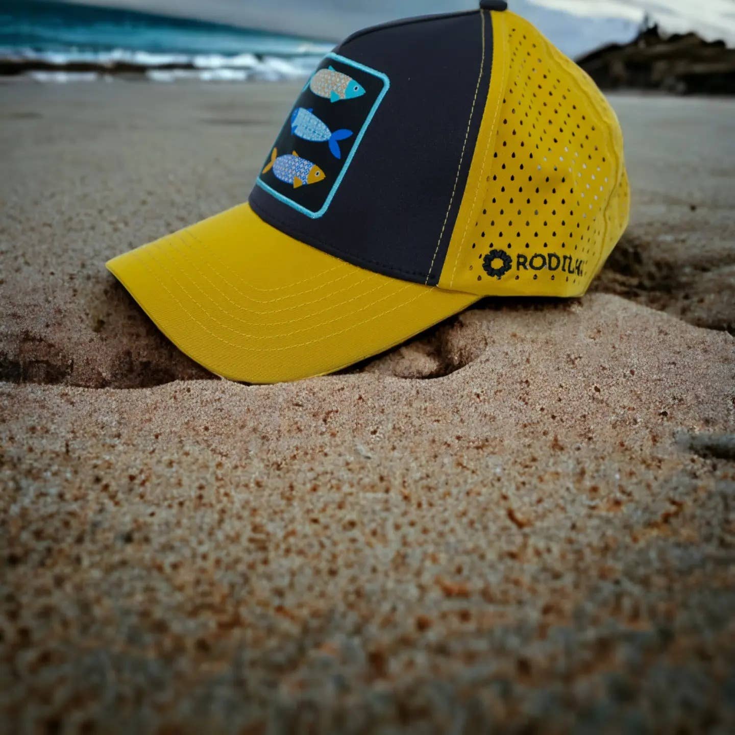 A Cap-Sardines and Tiles (Yellow and Black) by Rodilha, featuring fish illustrations, is partially buried in sand on a beach near the shoreline with ocean waves in the background.