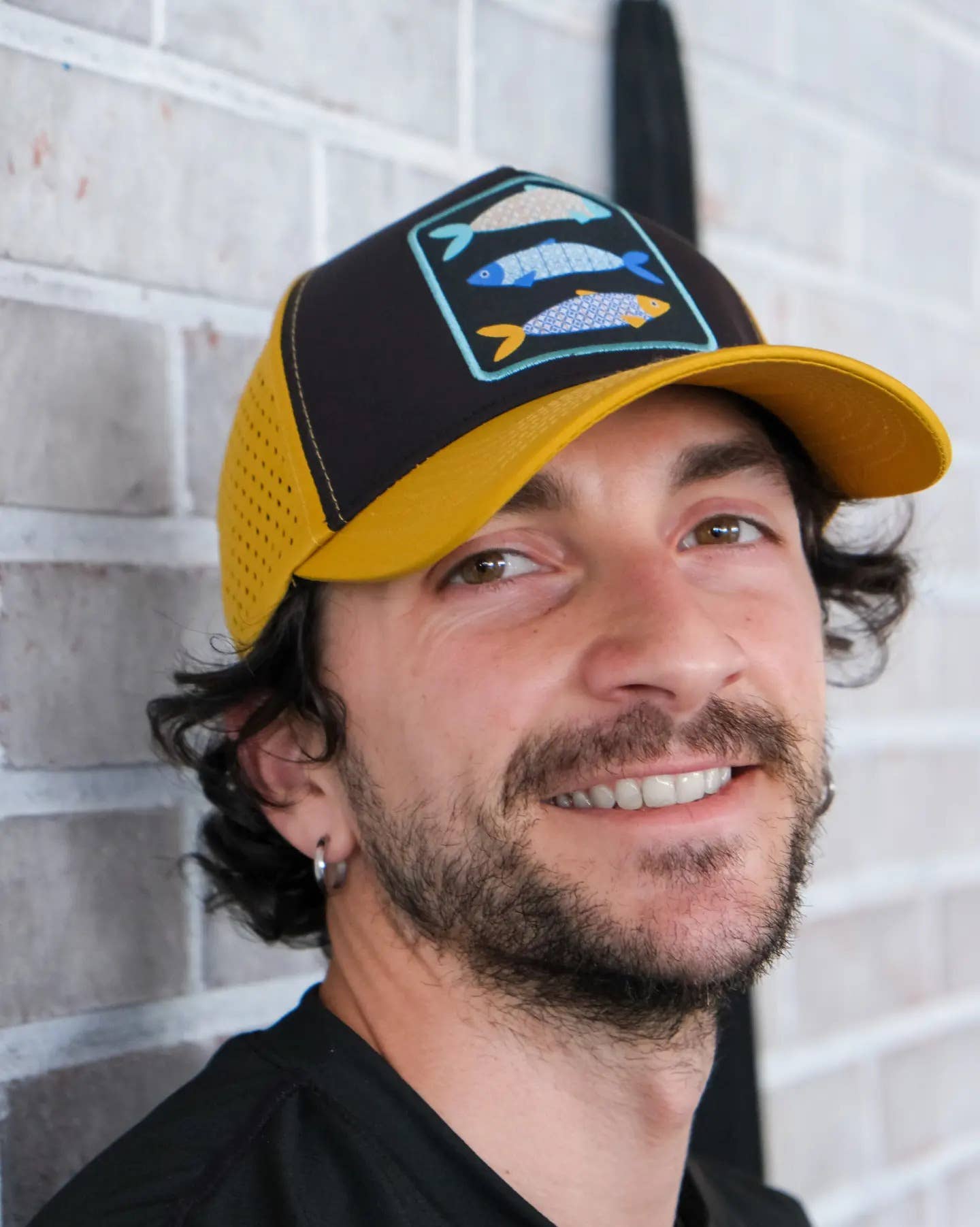 A man with wavy dark hair and a short beard smiles while wearing the Rodilha Cap-Sardines and Tiles (Yellow and Black), featuring three fish, as he stands before a light brick wall.