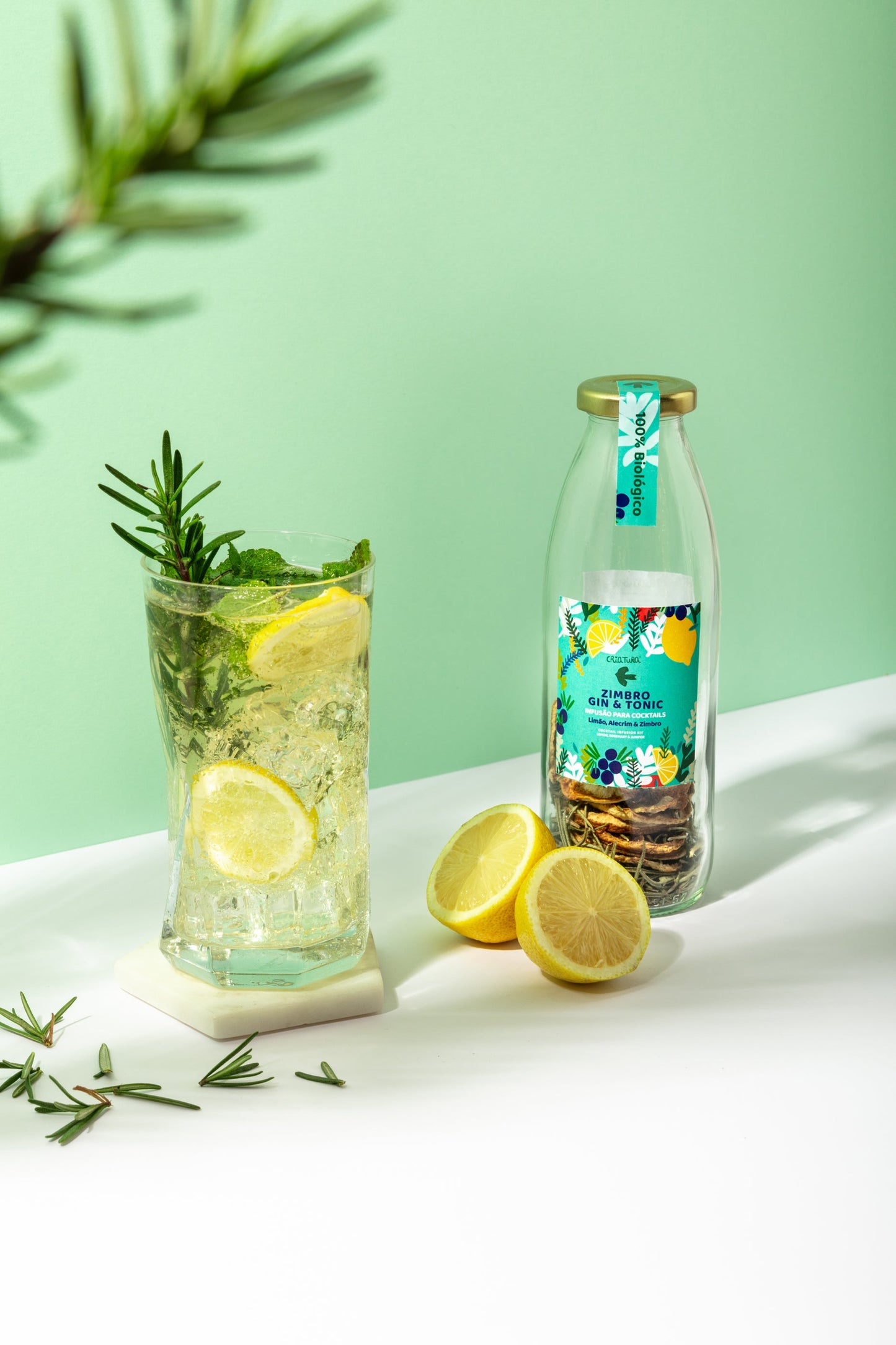 A glass of sparkling lemon drink with ice, rosemary, and juniper stands next to a bottle of Criatura Organic Cocktail Infusion – Lemon, Fennel & Juniper, fresh lemon halves, and rosemary sprigs on a white surface against a light green background.